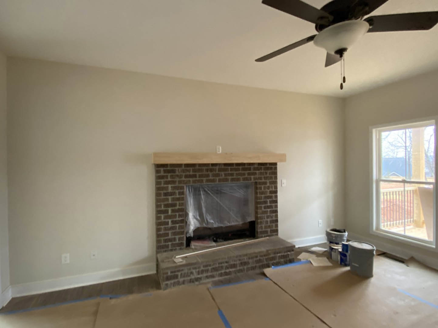 Living room with brick fireplace wrapped in plastic, ceiling fan with light fixture, window overlooking exterior railing, white storage container on floor, neutral walls and