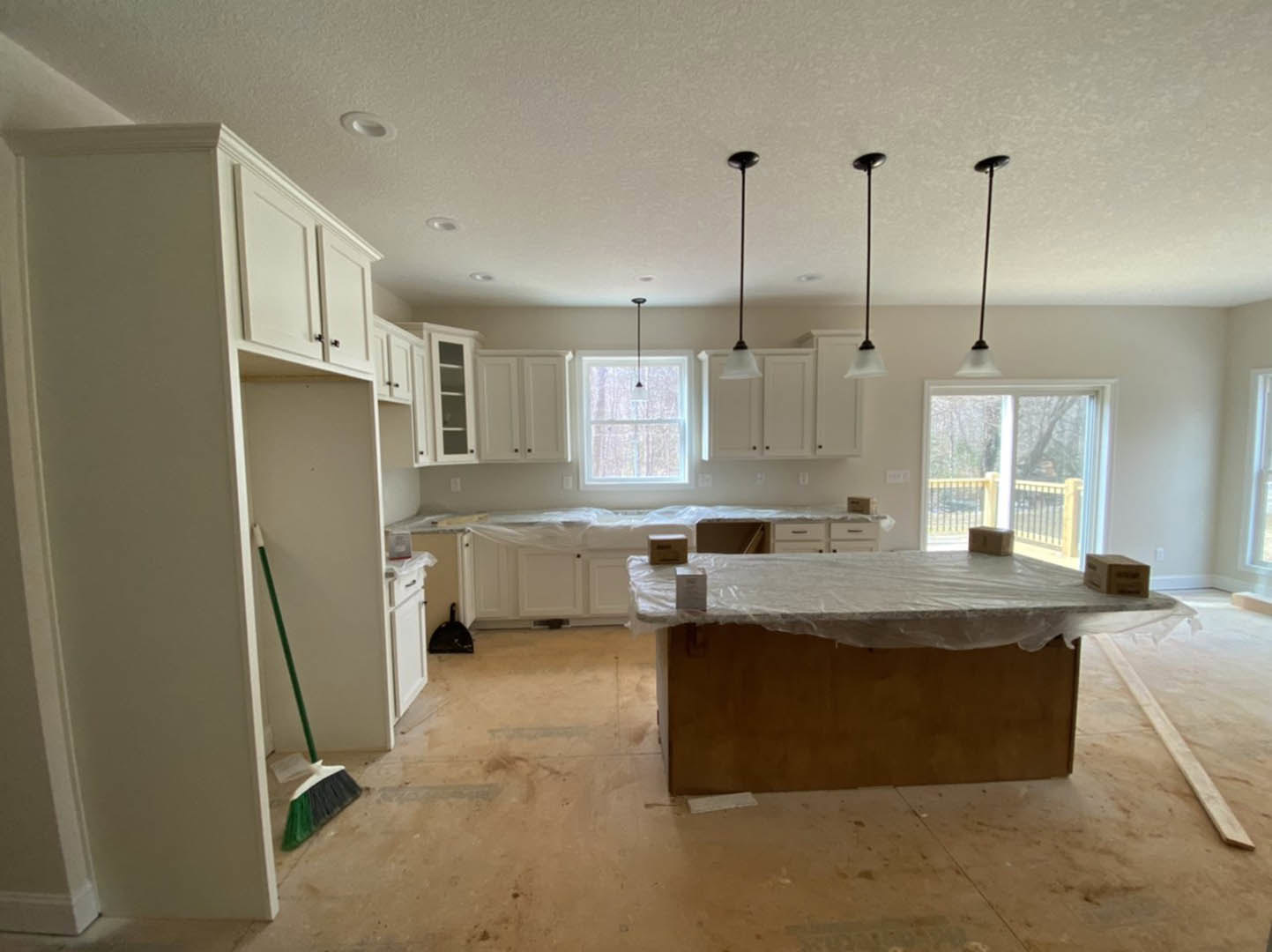 Modern kitchen with white cabinetry, wood table covered in plastic, broom leaning against wall, black dust pan on countertop, large window overlooking deck with storage box, row of