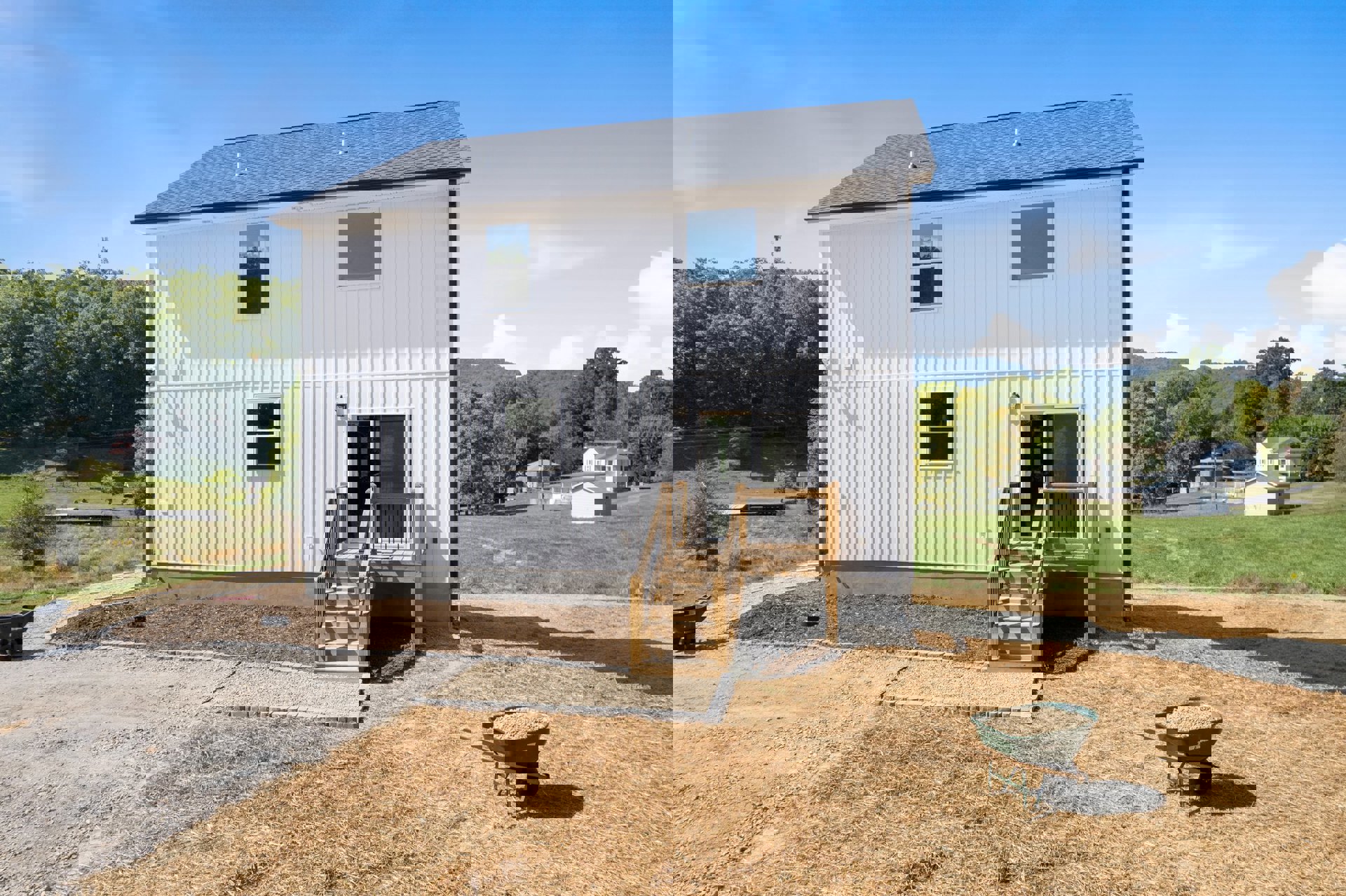White cottage-style house with white-framed windows, wooden staircase, wheelbarrow full of dirt on grassy yard, surrounded by trees and plants under a cloudy sky.