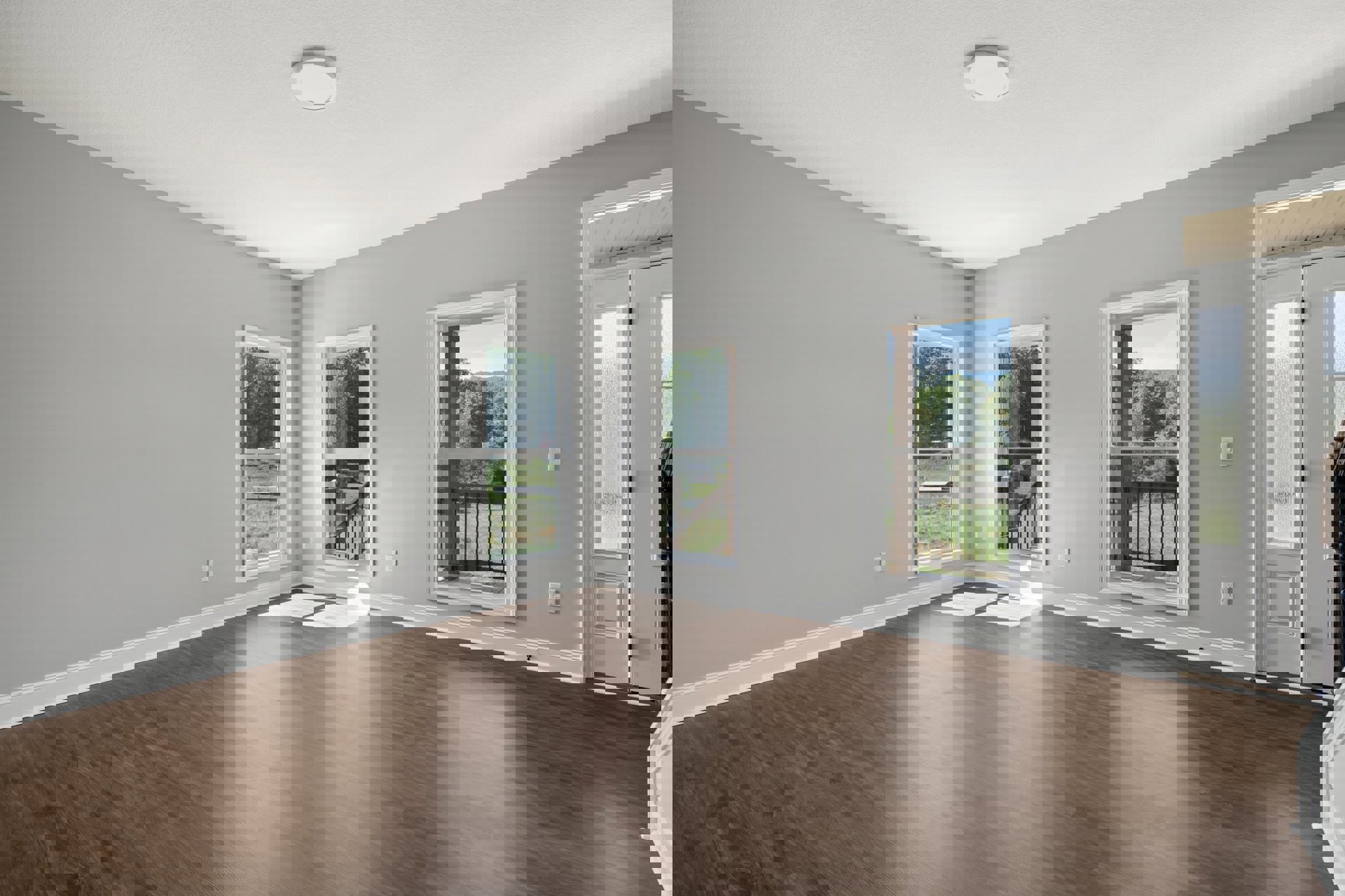 Wide-plank wood flooring in a bright room with large windows overlooking trees and grass, white walls and ceiling, minimal furnishings.