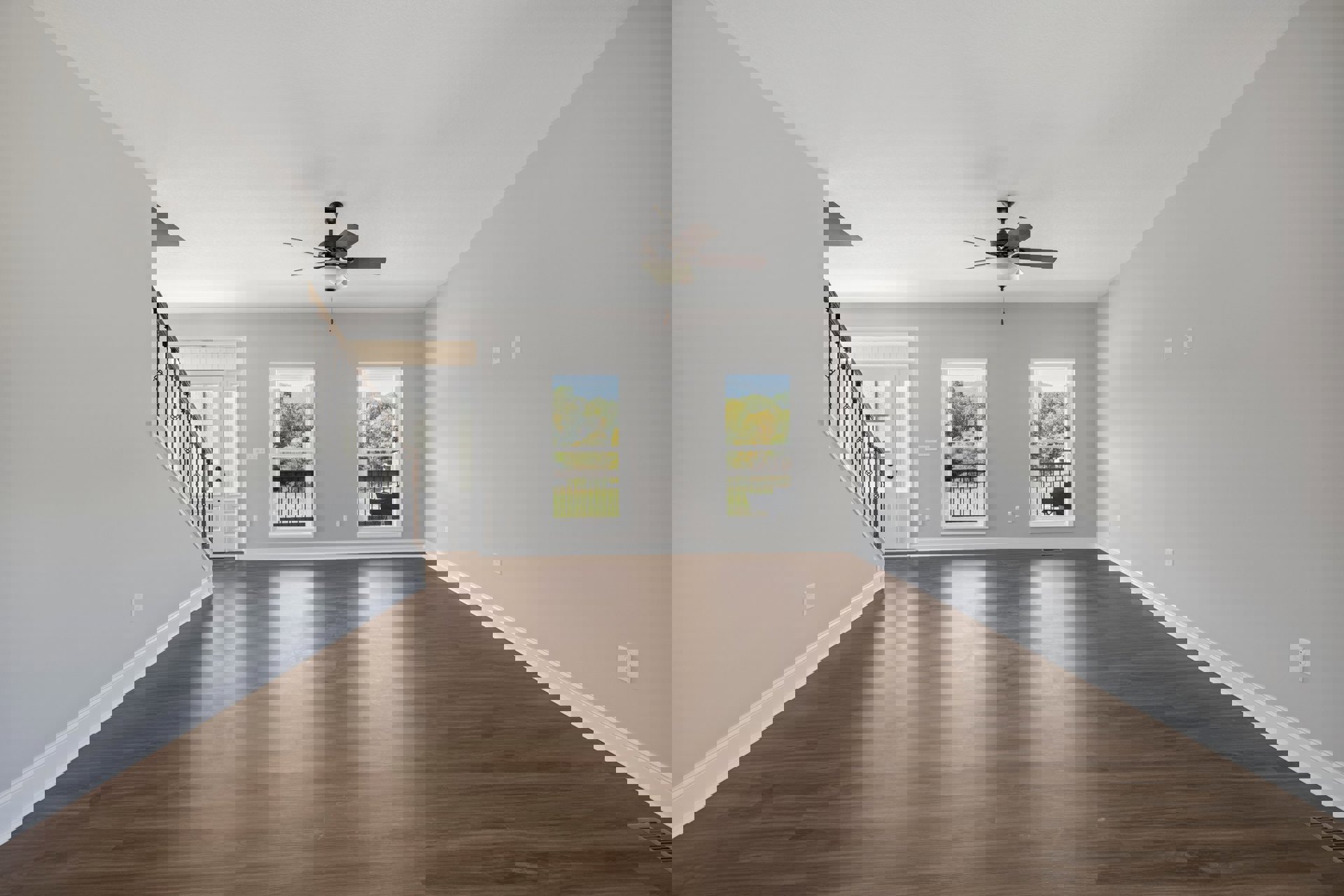 Spacious living area with hardwood floors, two windows overlooking trees, ceiling fan with light fixture, and wooden staircase leading to upper level; deck visible through window.