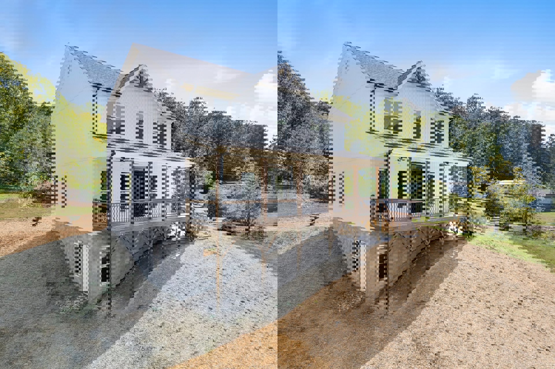 Two-story house with wood siding, large windows, and a spacious deck featuring black metal railing, situated on a grassy hill with trees in the background.