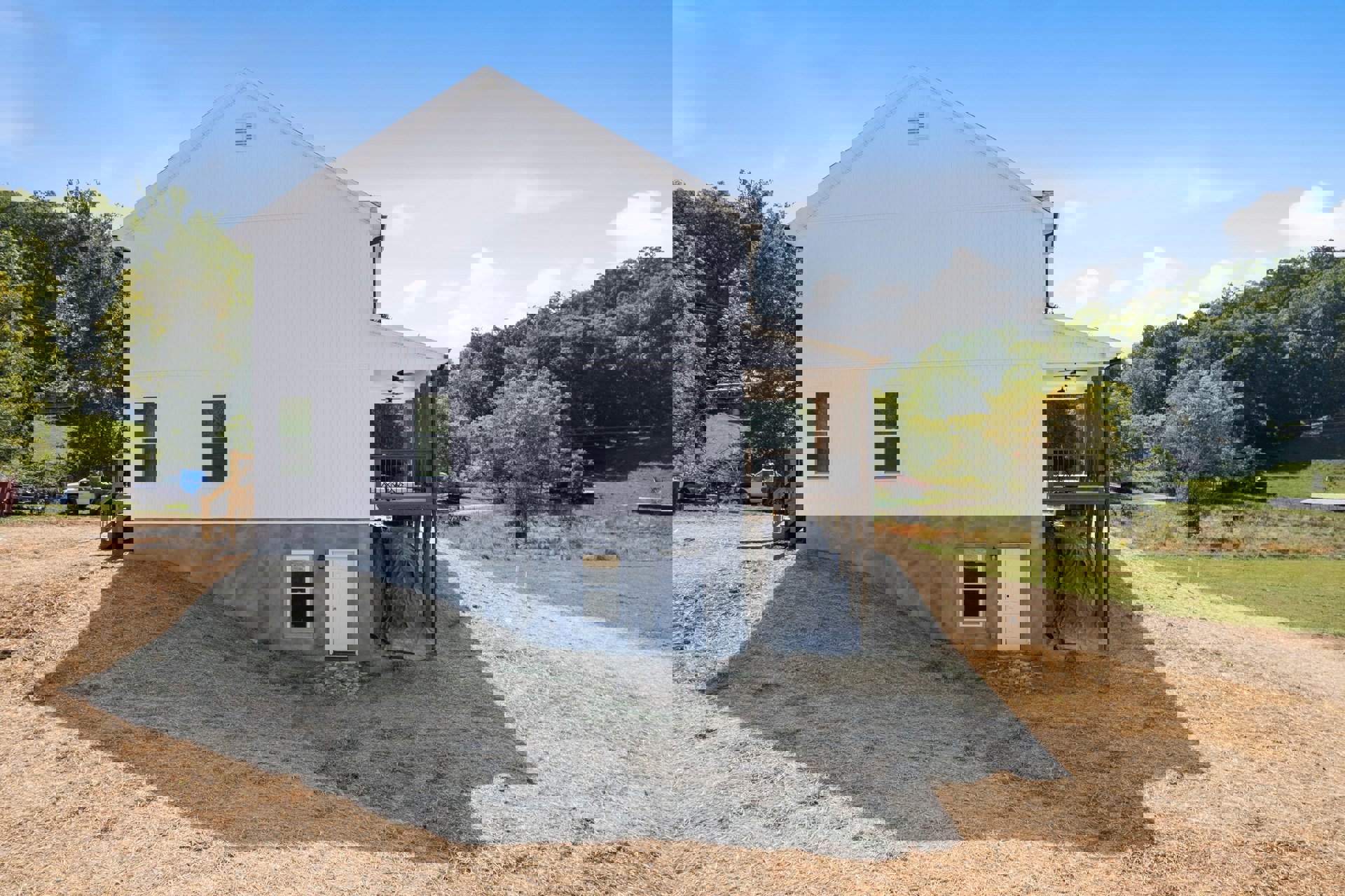 Partially built house with exposed framing, white window and door, black tarp covering concrete foundation, wooden deck and porch, grassy lot bordered by trees in background