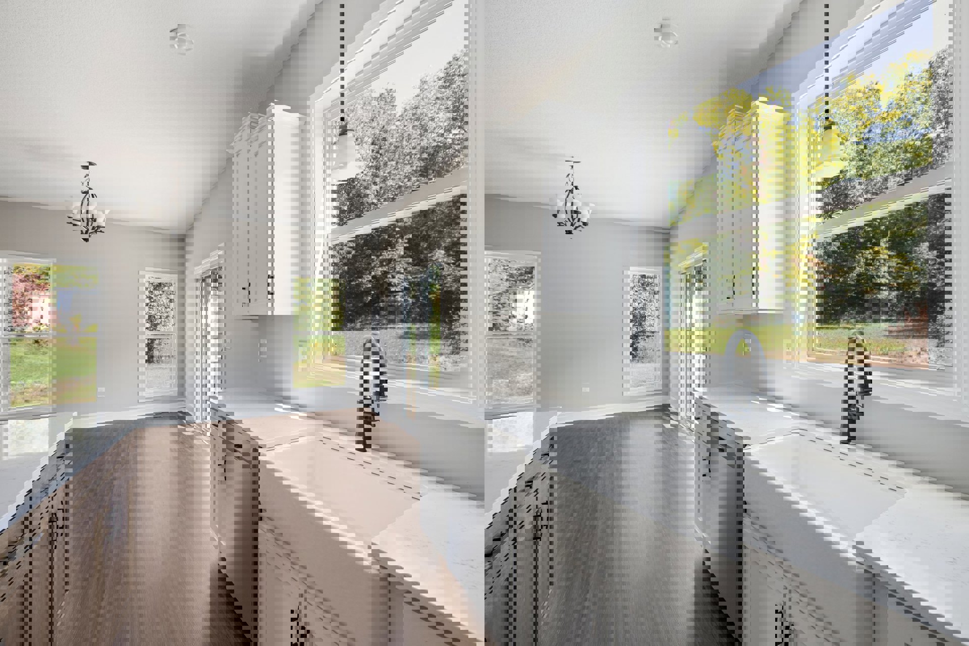 White kitchen cabinets with silver hardware, wood plank flooring, black faucet at a white farmhouse sink, large window overlooking trees and neighboring house, quartz countertops