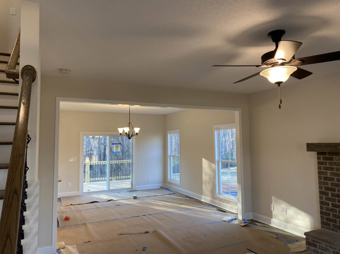 Ceiling fan with integrated light fixture mounted on white plaster ceiling in a spacious room, wooden staircase post visible in foreground, window with sign in background, close-up
