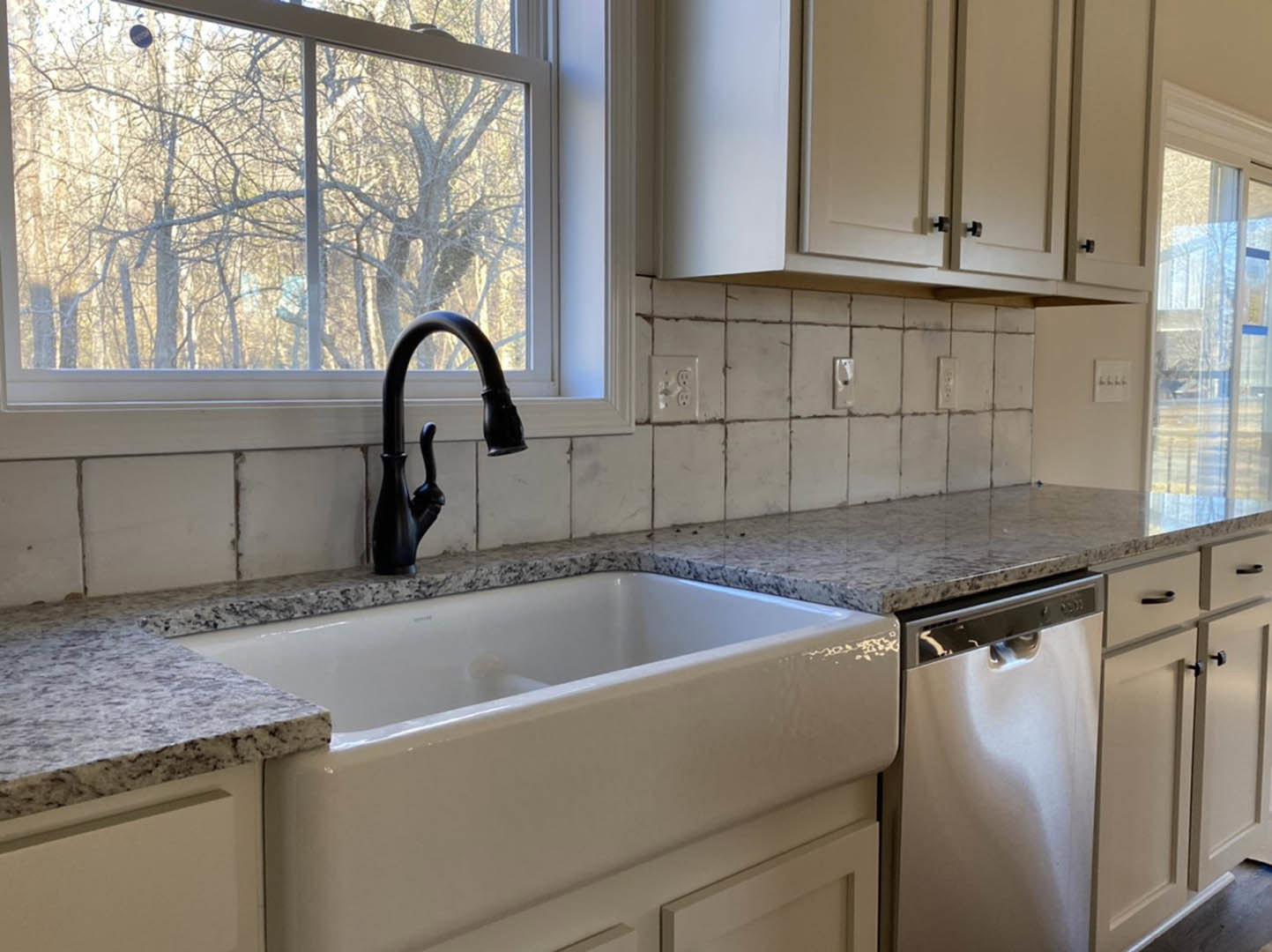 Black kitchen faucet and stainless sink set in marble countertop, white tile backsplash, window with trees outside, white cabinetry, dishwasher, three-hook light switch, close-up