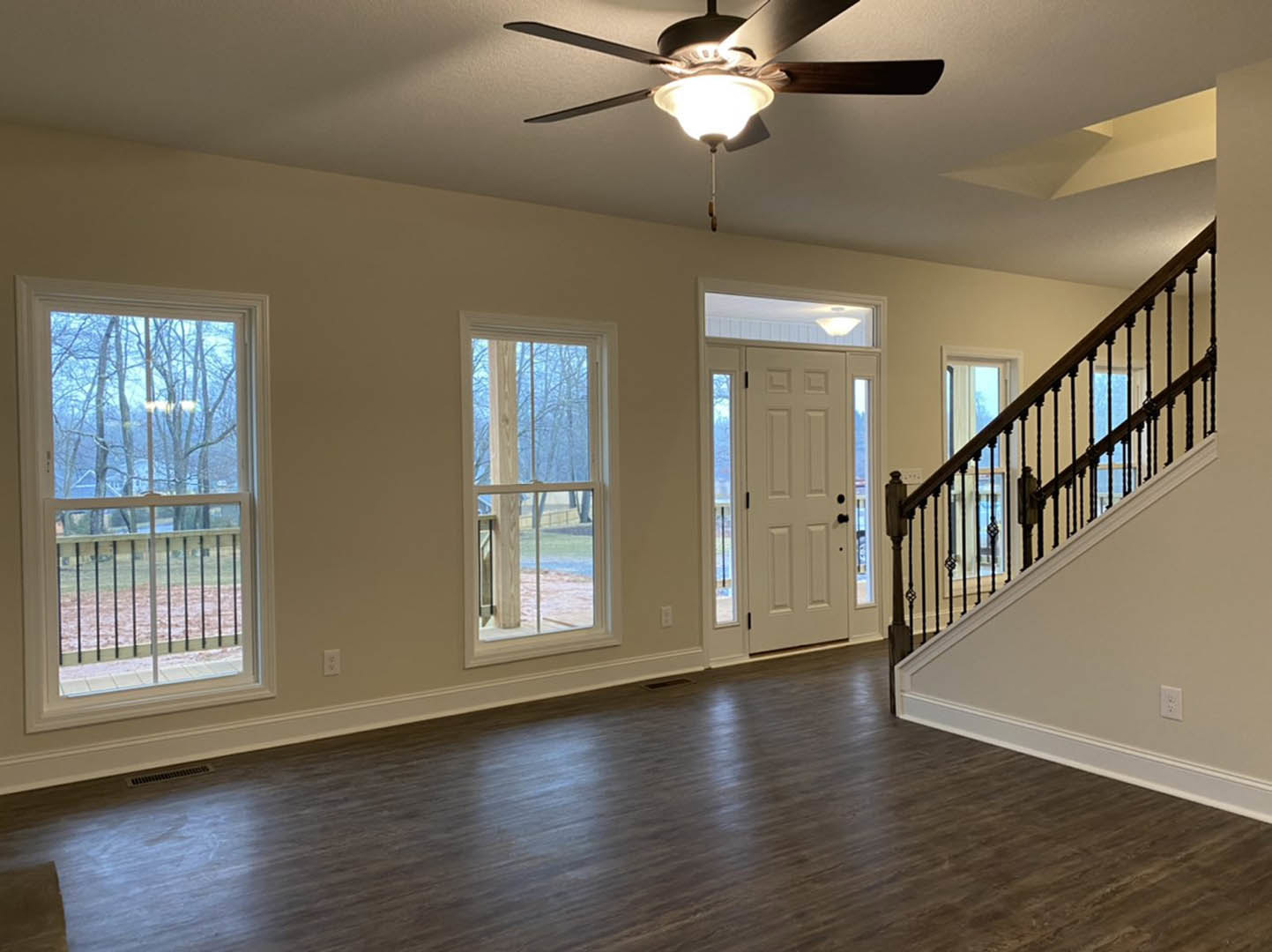 Open living area with hardwood floors, black metal staircase railing, large window overlooking trees, and ceiling fan with light fixture