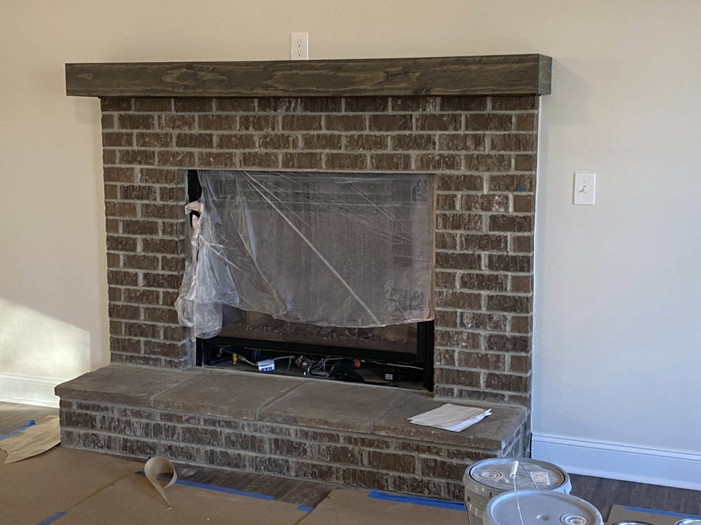 Brick fireplace with wood mantle, plastic covering over hearth, wood floor, light switch on adjacent wall, close-up of brick step and paper nearby