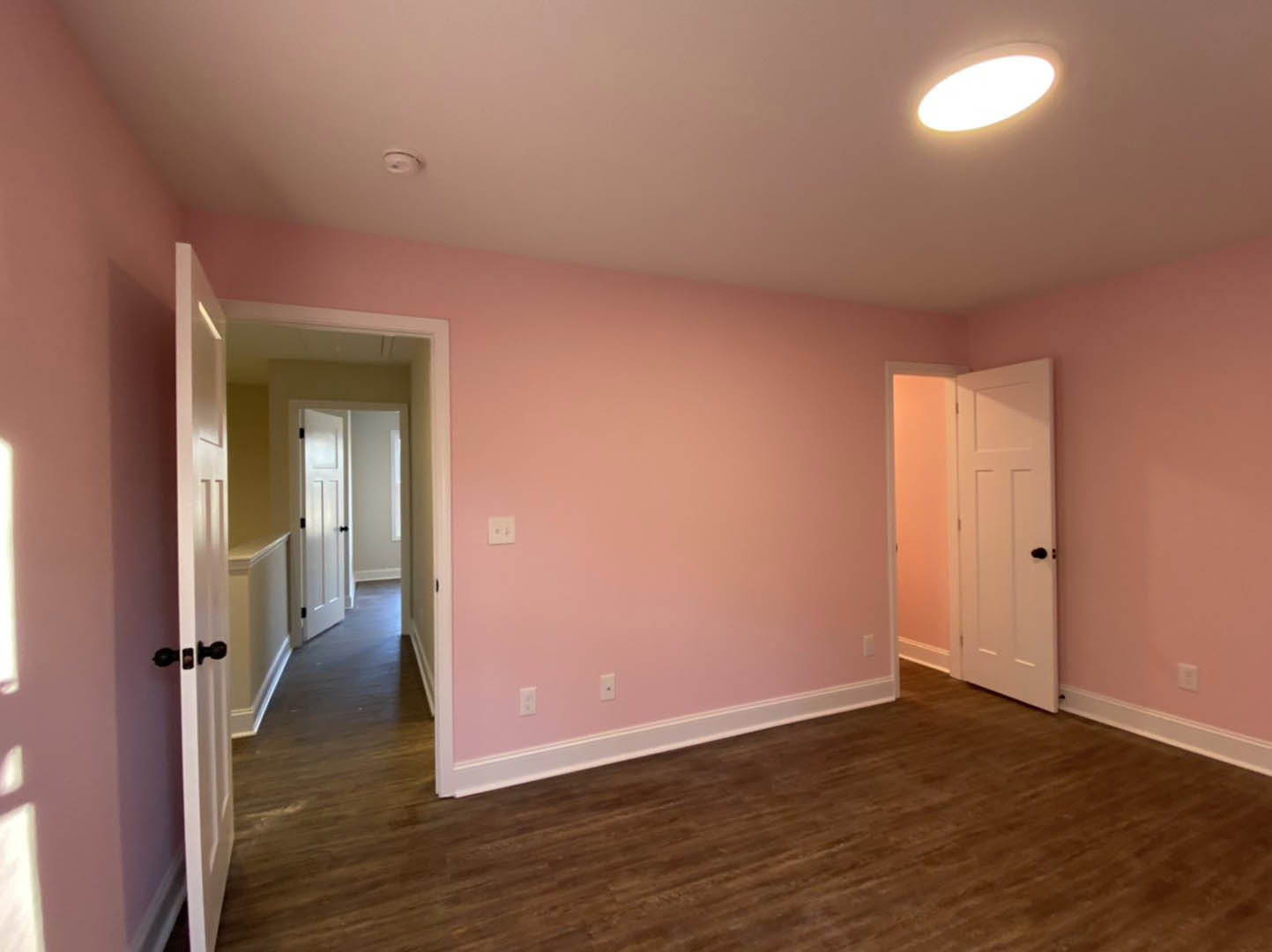 Pink-painted walls and wood laminate flooring in a bright room, white door with black handle, ceiling light fixture, hallway with additional white doors visible.