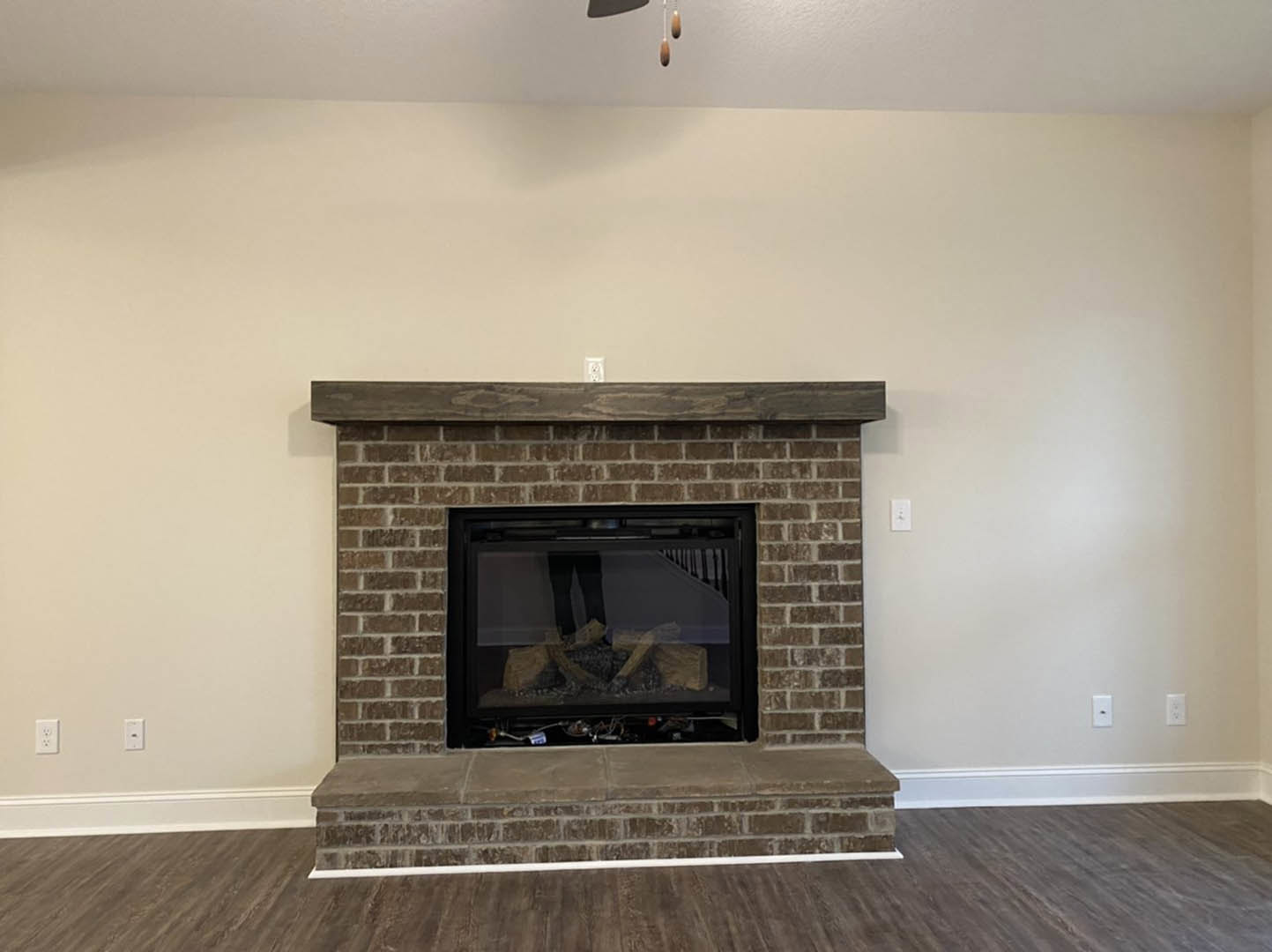 Brick fireplace with stacked wood logs, wood floor, white trim, and built-in shelf against a neutral wall in a cozy living room.