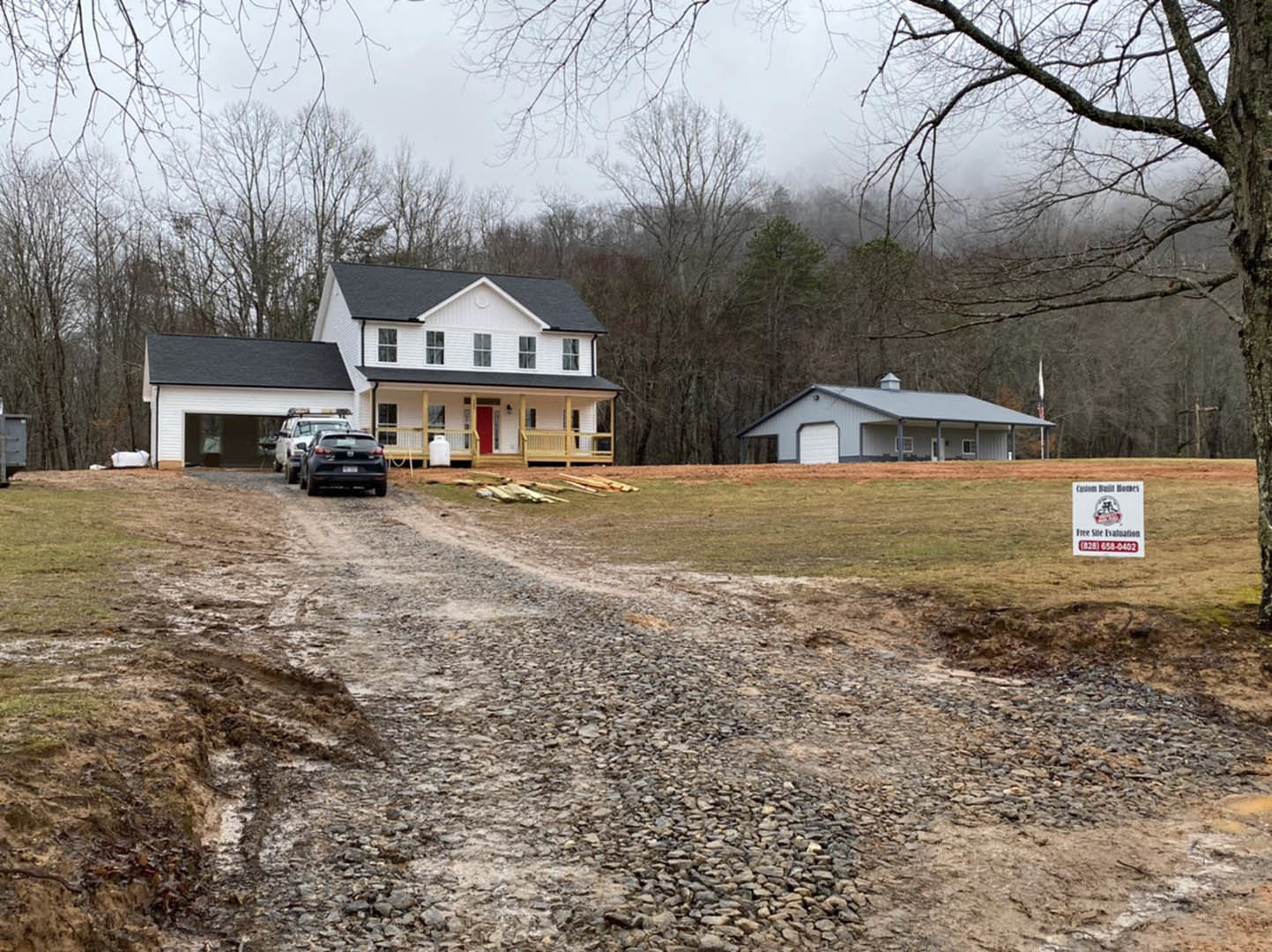 Modern house with light-colored siding, dark roof, gravel driveway, parked black car, grassy lawn, white sign with red and black text, mature trees in background