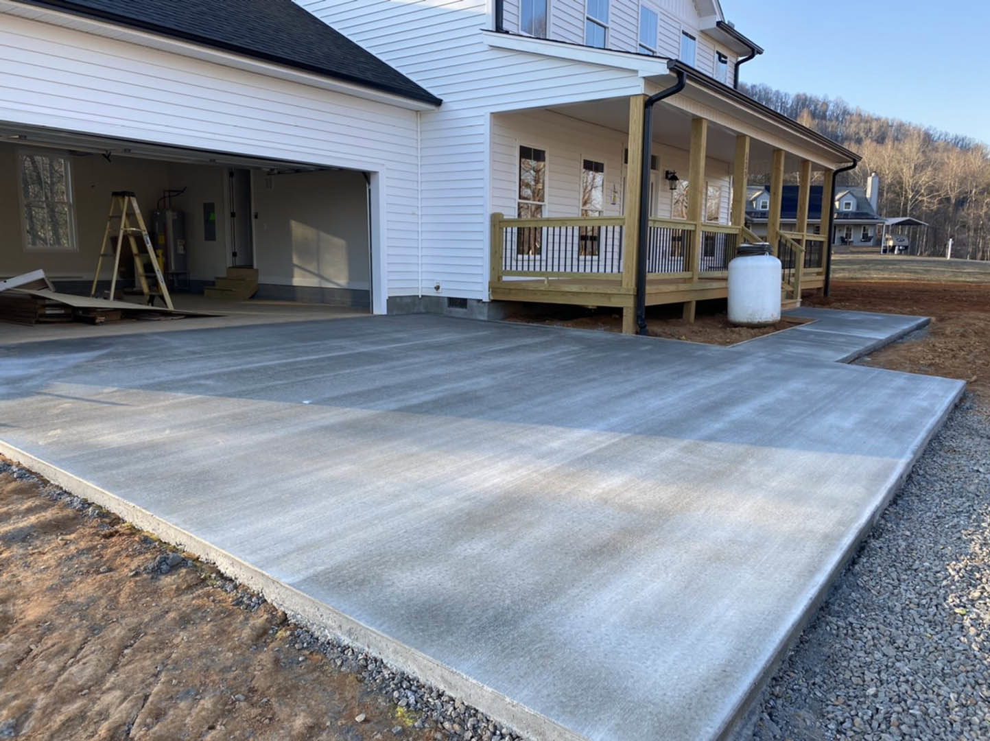Concrete driveway leading to a modern home with large windows, white exterior walls, and a ladder propped against the porch; trees and sky visible in the background.