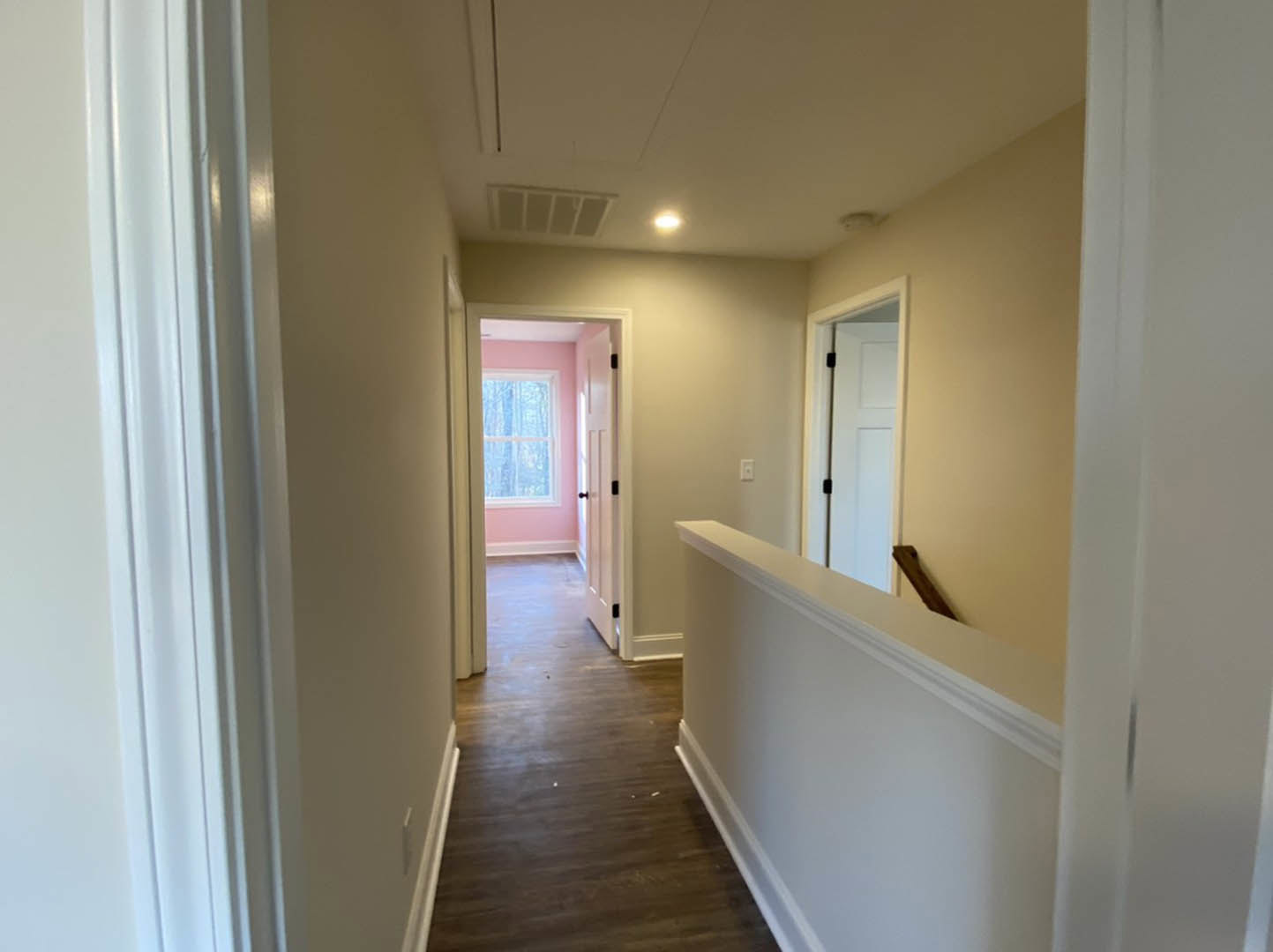 Hallway with wood flooring, white railing, pink door, white walls, and window with white frame