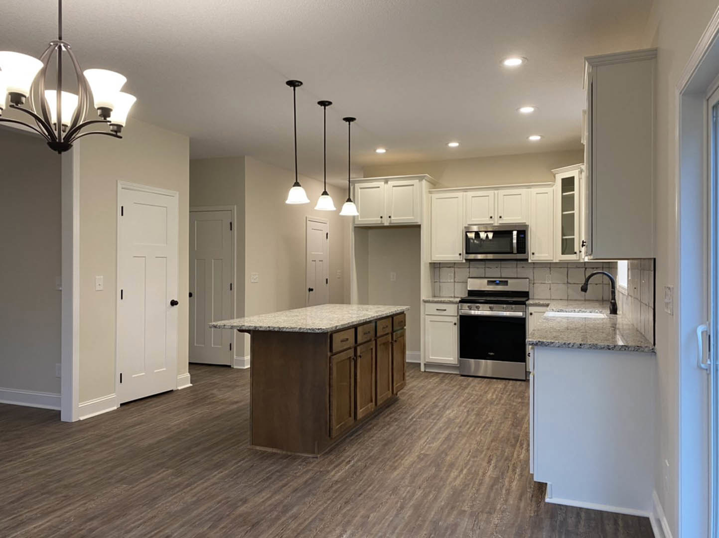 Kitchen with light wood flooring, white shaker cabinets, marble-topped island, stainless steel stove and microwave, modern chandelier, and white door with black hardware
