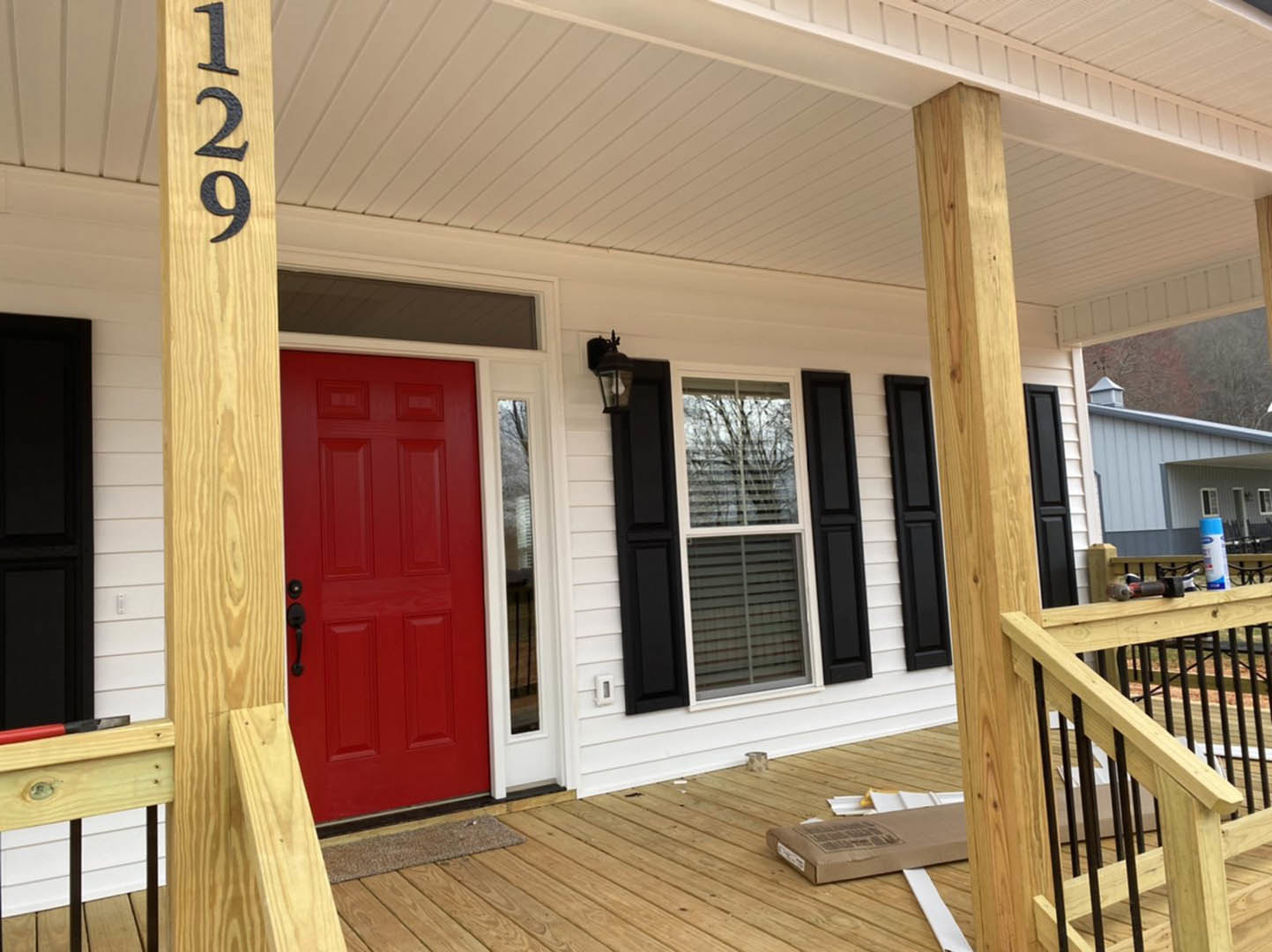Red front door framed by wooden pillars and black metal railings on a porch, brown mailbox with newspaper, house number mounted on wood siding, wood deck flooring.