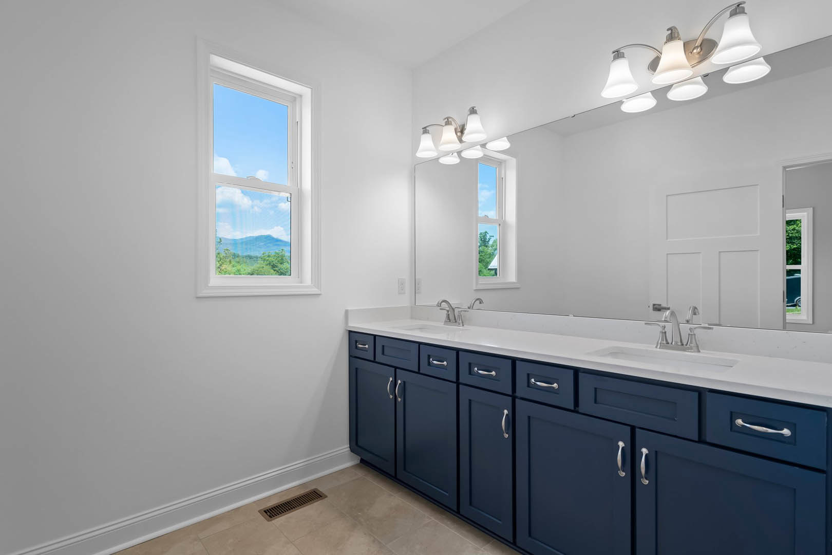 Bathroom with expansive wall mirror, blue shaker-style cabinets, white countertop, chrome faucet, tile flooring, window showing blue sky, row of modern light fixtures above mirror