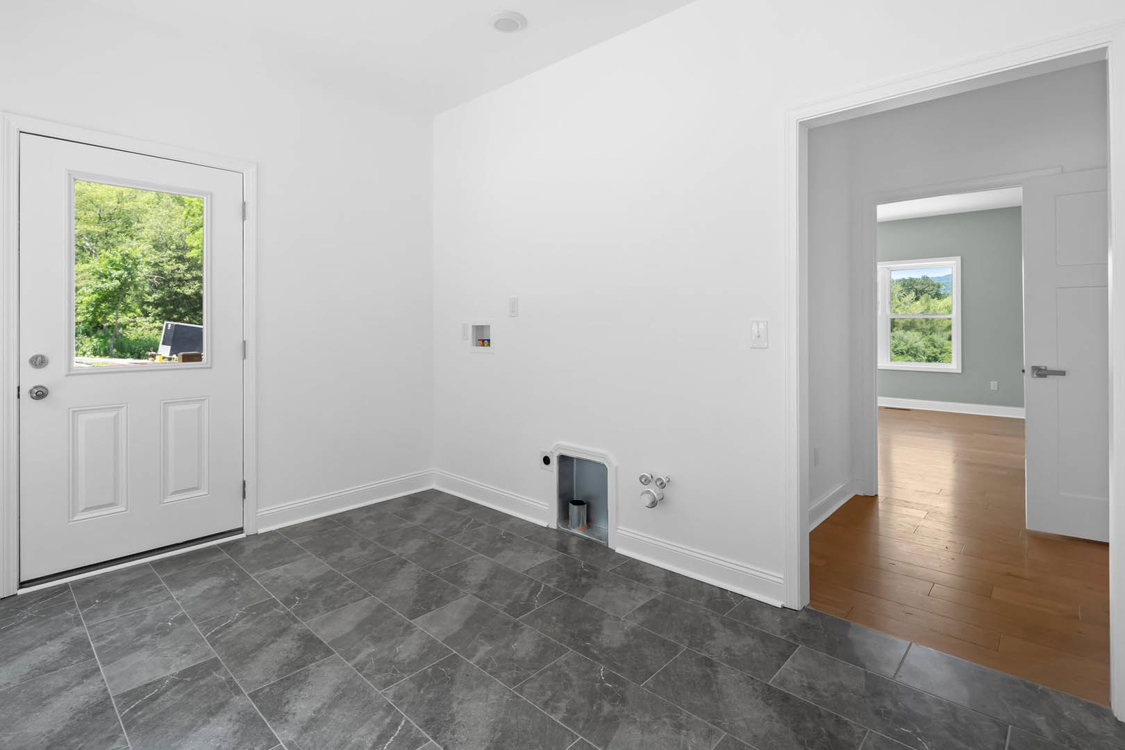 White door with glass window, grey tile floor, white walls, window overlooking green trees, group of silver knobs on white surface, hole in wall near floor.