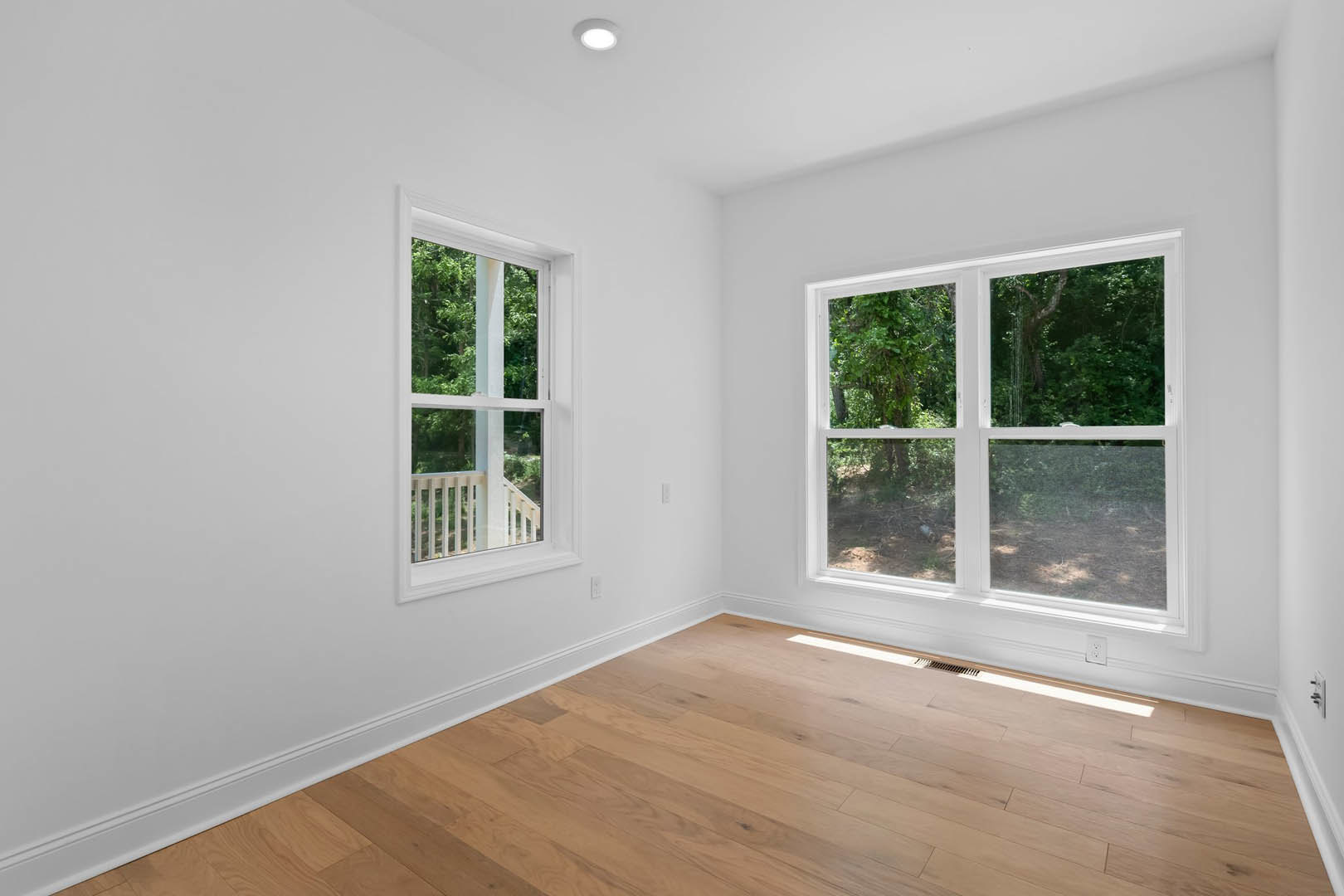Spacious room featuring large windows with views of leafy trees, natural light streaming onto light wood flooring, white walls, ceiling light fixture, and a person walking along a