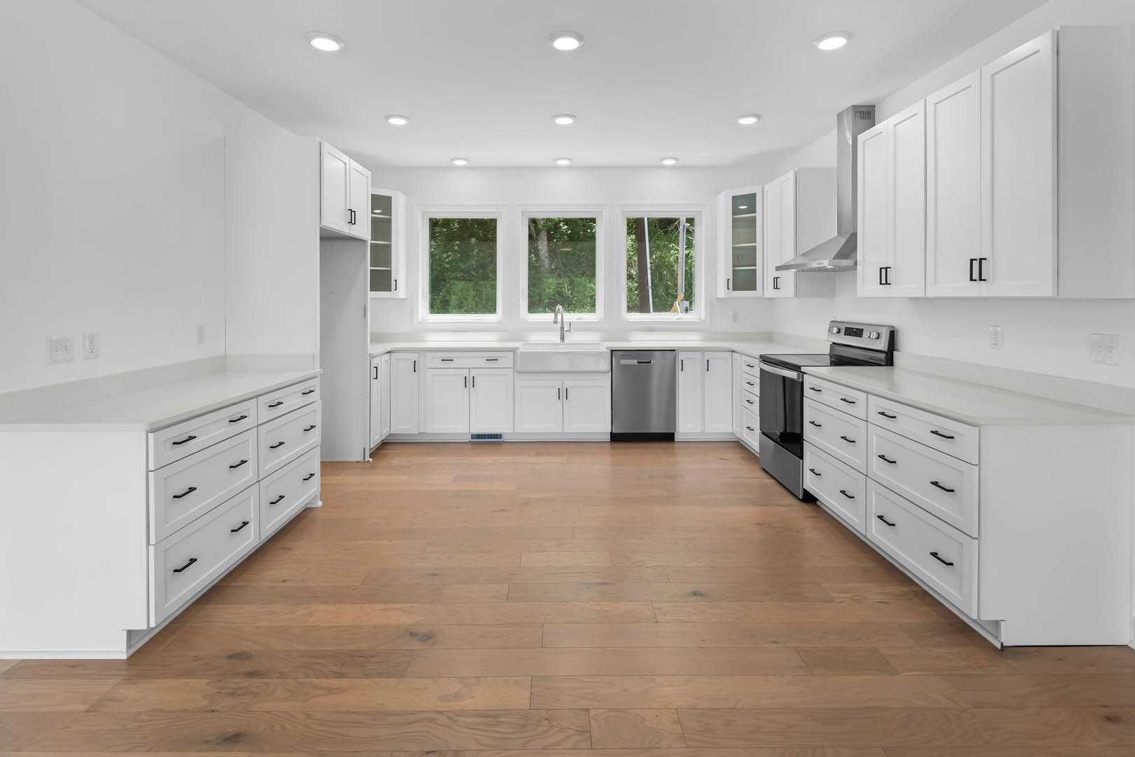White shaker cabinets, stainless steel refrigerator, wood plank flooring, quartz countertops, large window with white trim overlooking trees and bushes.