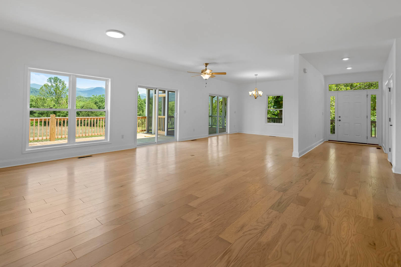 Spacious room with hardwood flooring, multiple windows, ceiling fan with light, white door featuring black handle, and view of wooden deck with white post