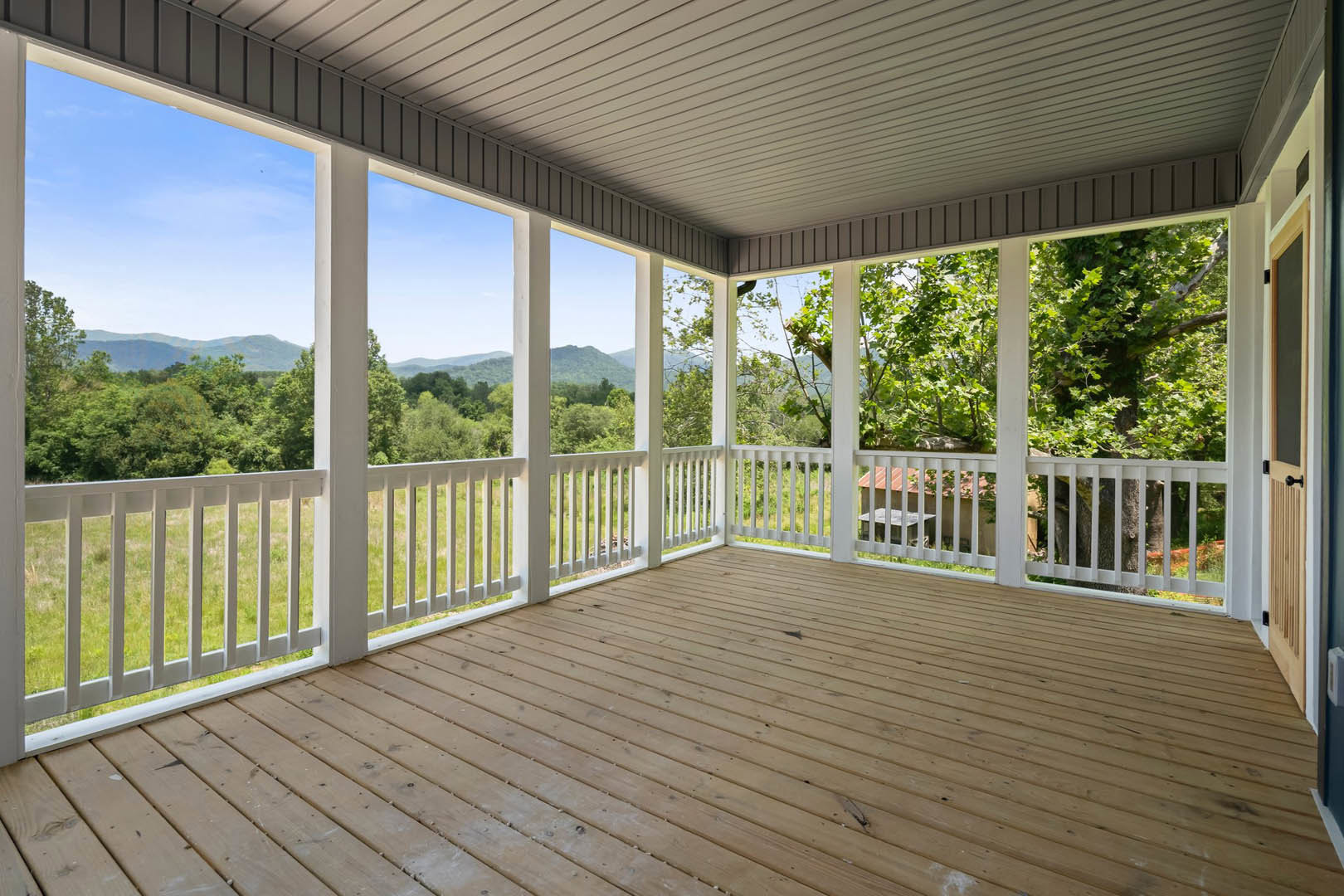 Spacious wooden porch with white railings, white ceiling with black trim, overlooking green trees and distant mountains under a clear blue sky