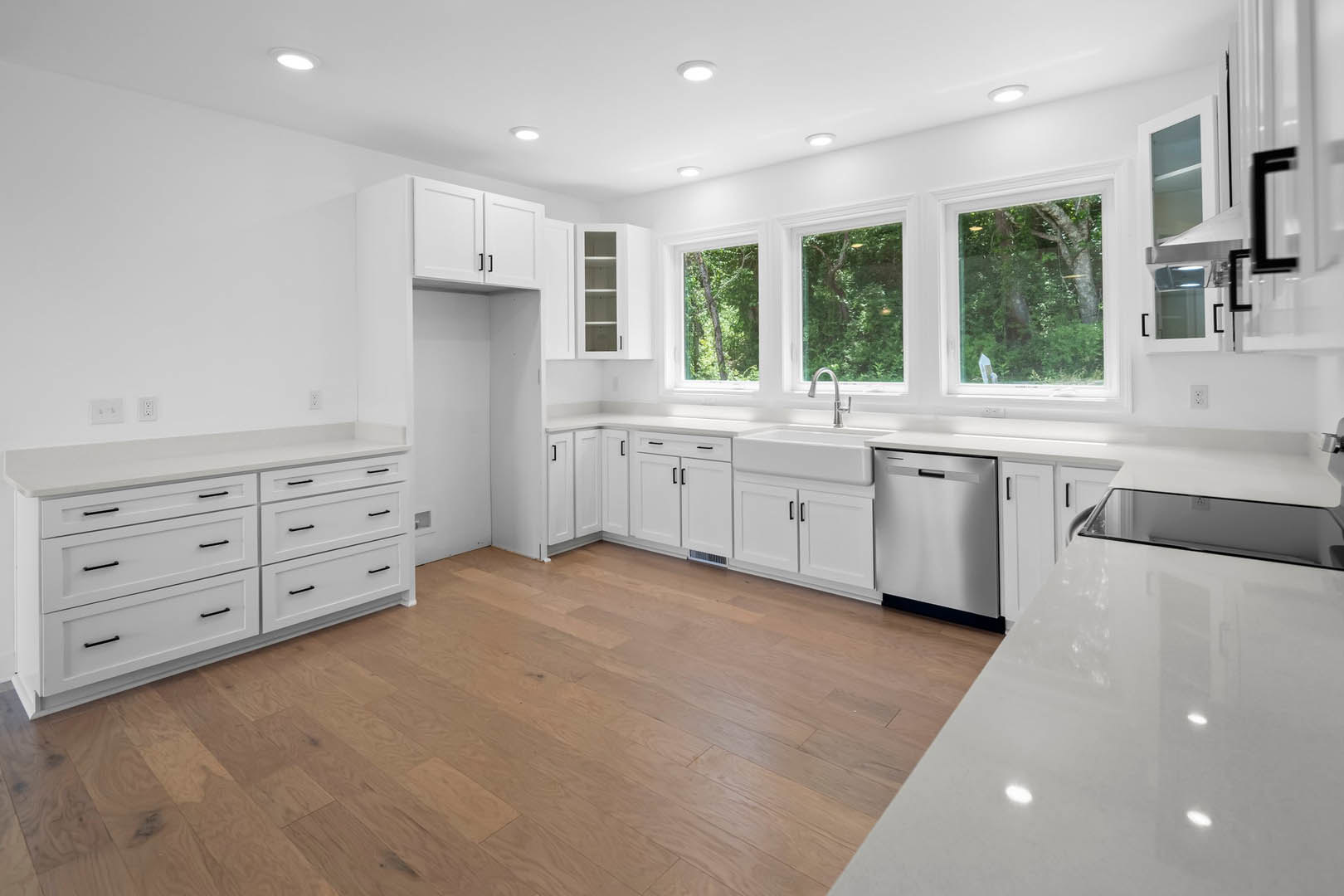 White kitchen cabinets with black handles, white countertop, stainless steel dishwasher, hardwood flooring, window overlooking a leafy tree.