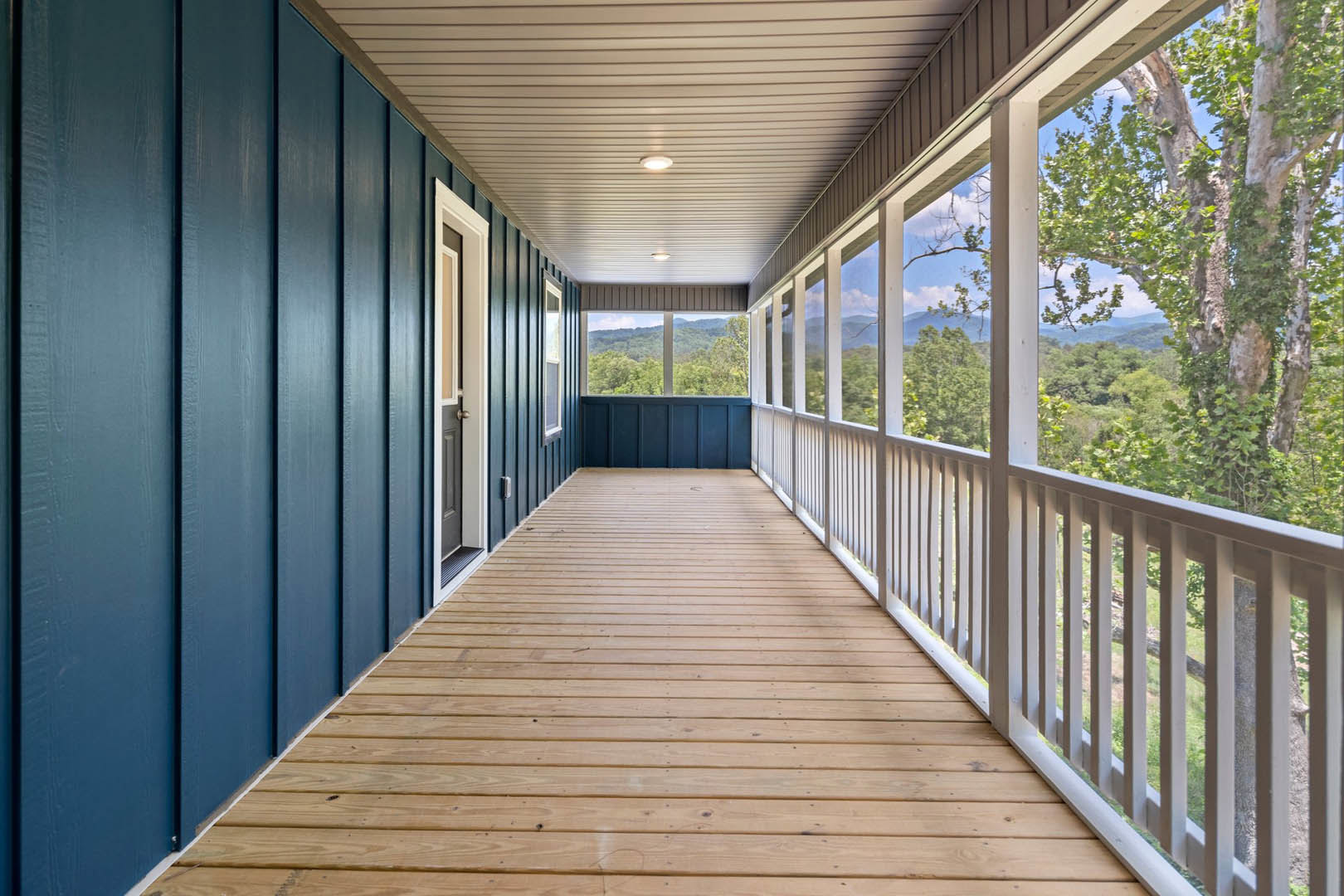 Wood deck with white railing, wood floor, and door opening to porch; surrounded by green trees and mountain views; ceiling features recessed lights, window visible on exterior