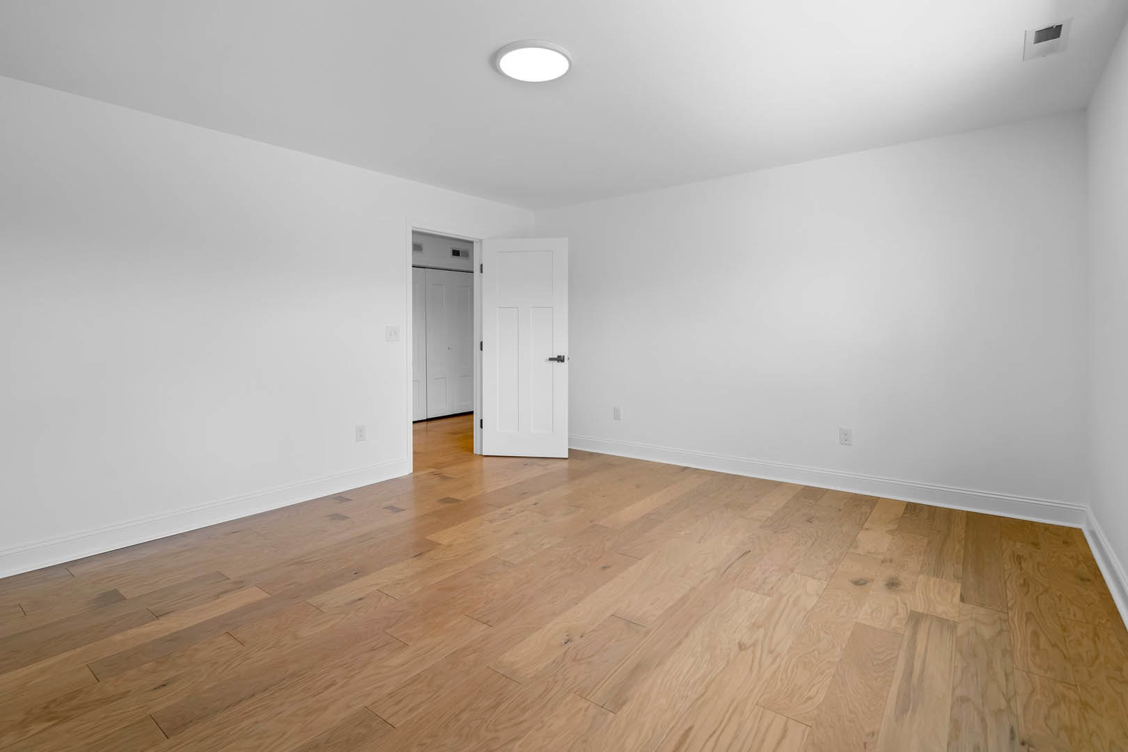 White paneled door with black handle open onto hardwood floor, white walls, ceiling light, wall vent, and black wall-mounted object.