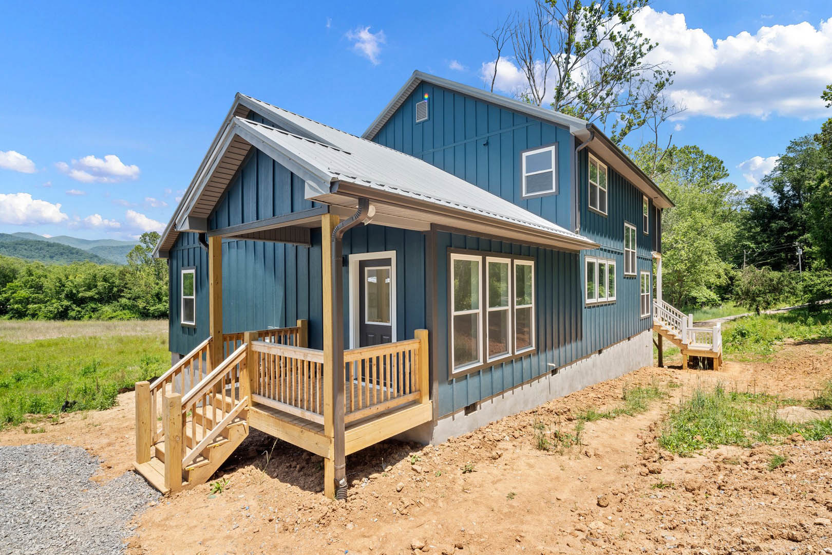 Two-story house with wooden porch and staircase, white siding, large windows, wooden deck with railing, front door, tree in yard, blue sky with clouds