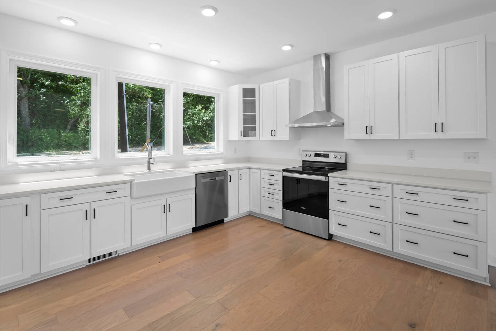 White shaker cabinets, stainless steel stove, hardwood flooring, quartz countertops, window overlooking trees, built-in dishwasher, brushed metal support pole