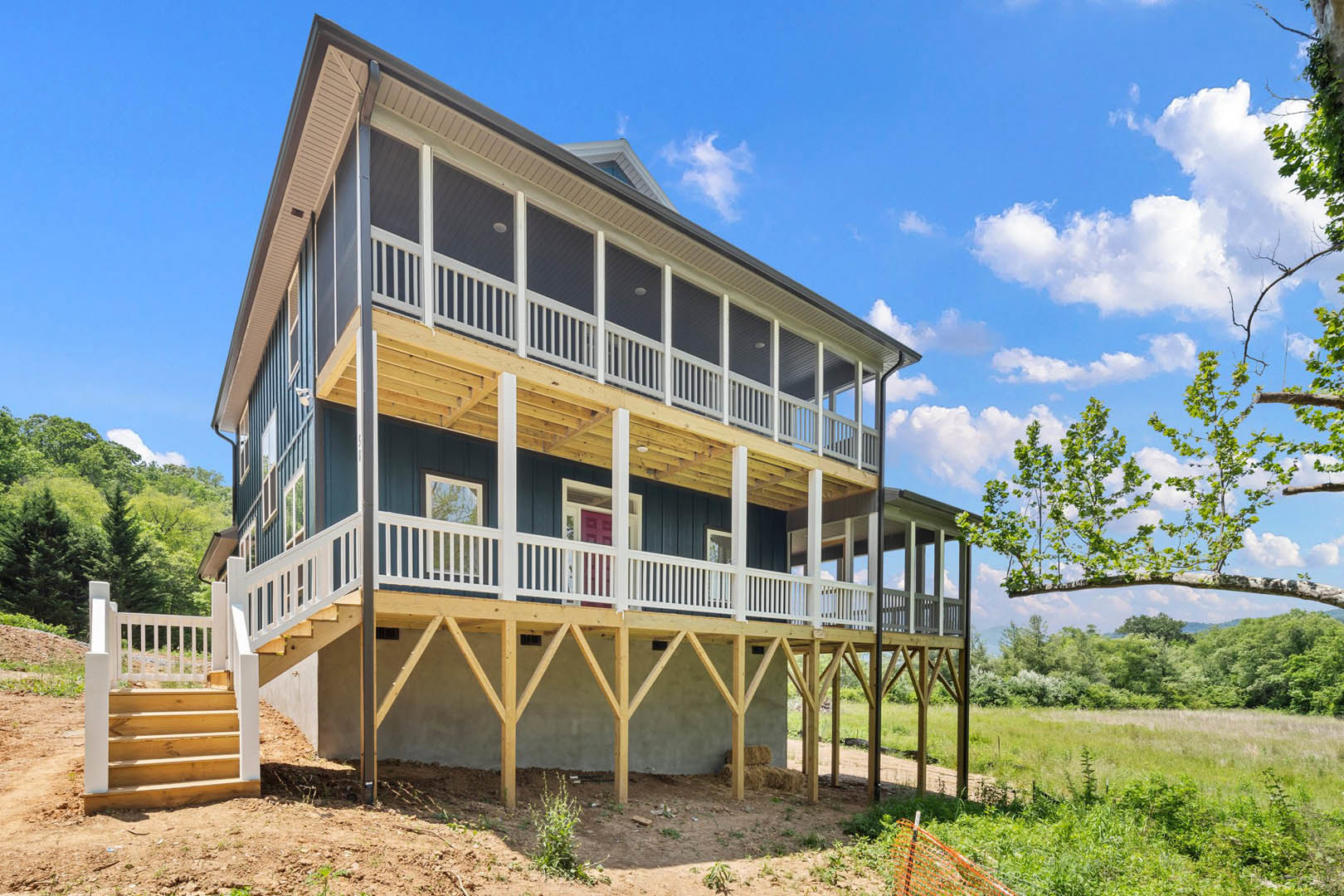 Two-story home with wide covered porch, wooden steps, white railing, large windows, surrounded by mature trees and landscaped yard