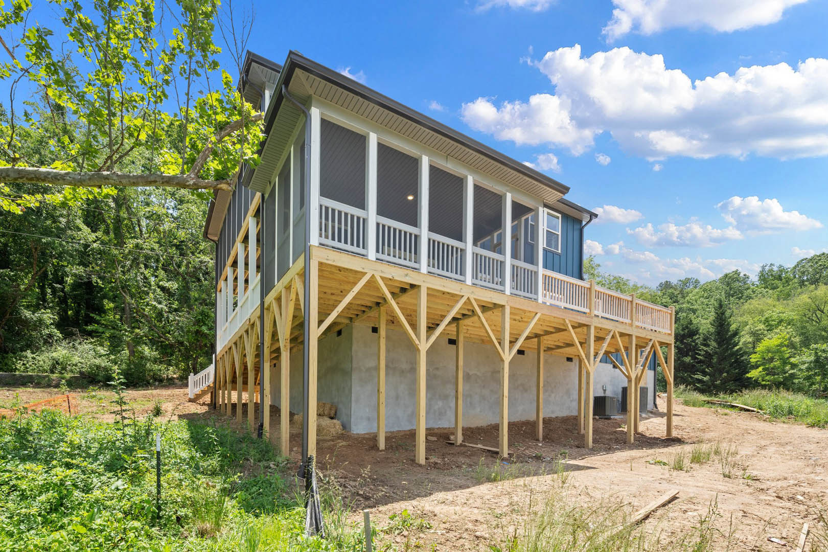 Two-story house with white siding, covered front porch, wooden deck, large windows, mature tree in landscaped yard, cloudy sky overhead