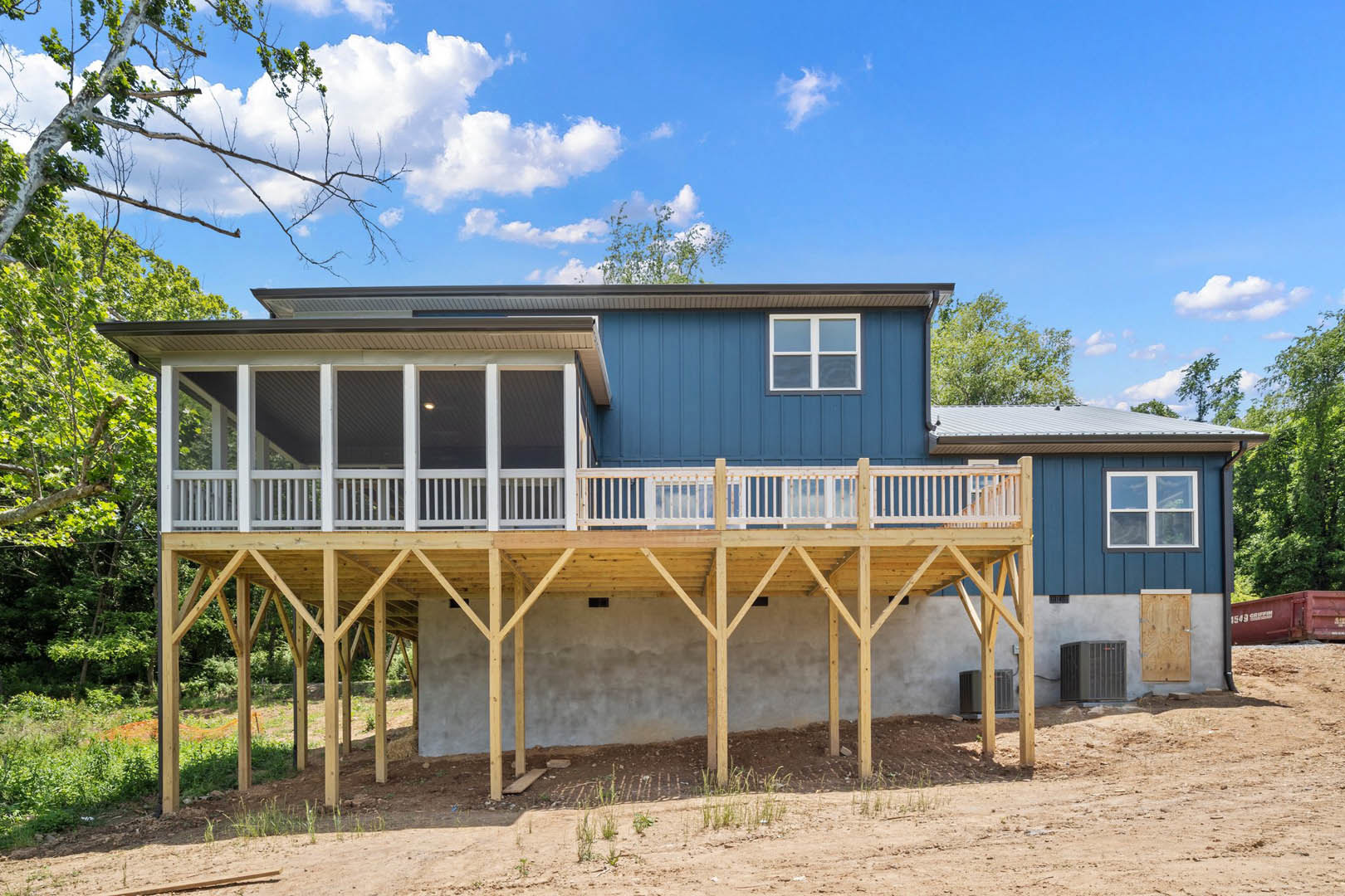 Wood deck with metal railing attached to a modern home, large windows with white trim, gray siding, surrounded by trees under a cloudy sky