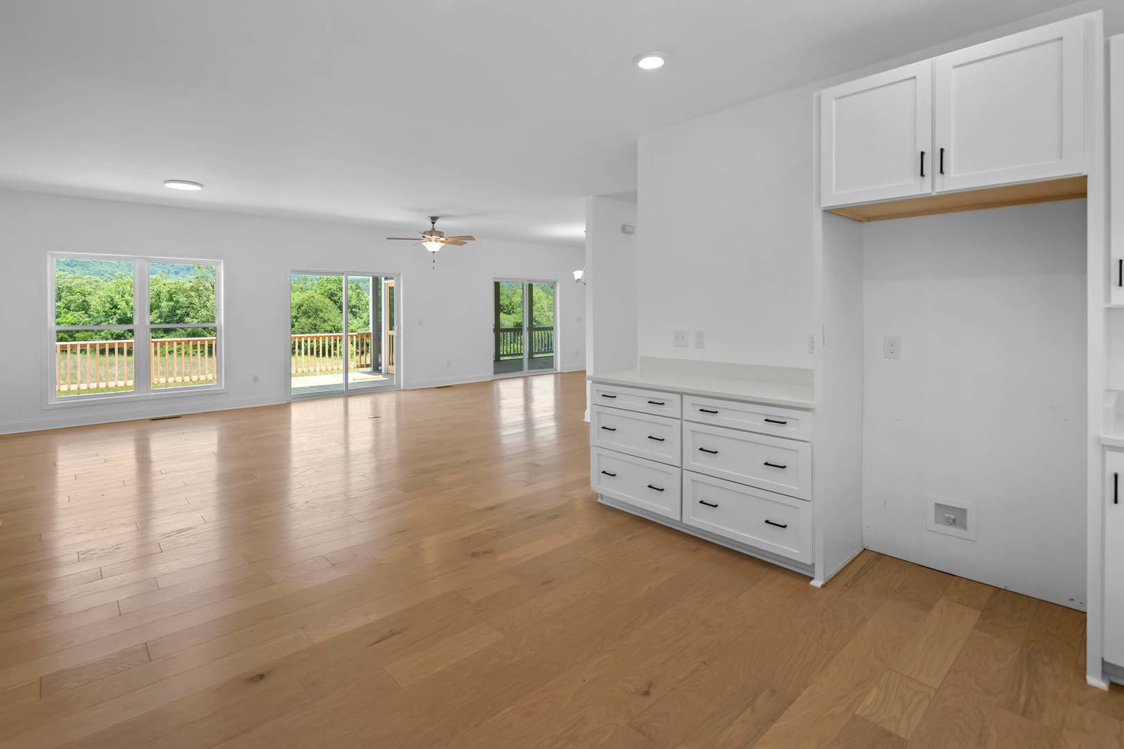 Spacious room featuring white cabinets with black handles, hardwood flooring, white walls, ceiling fan with light, large window overlooking trees and railing