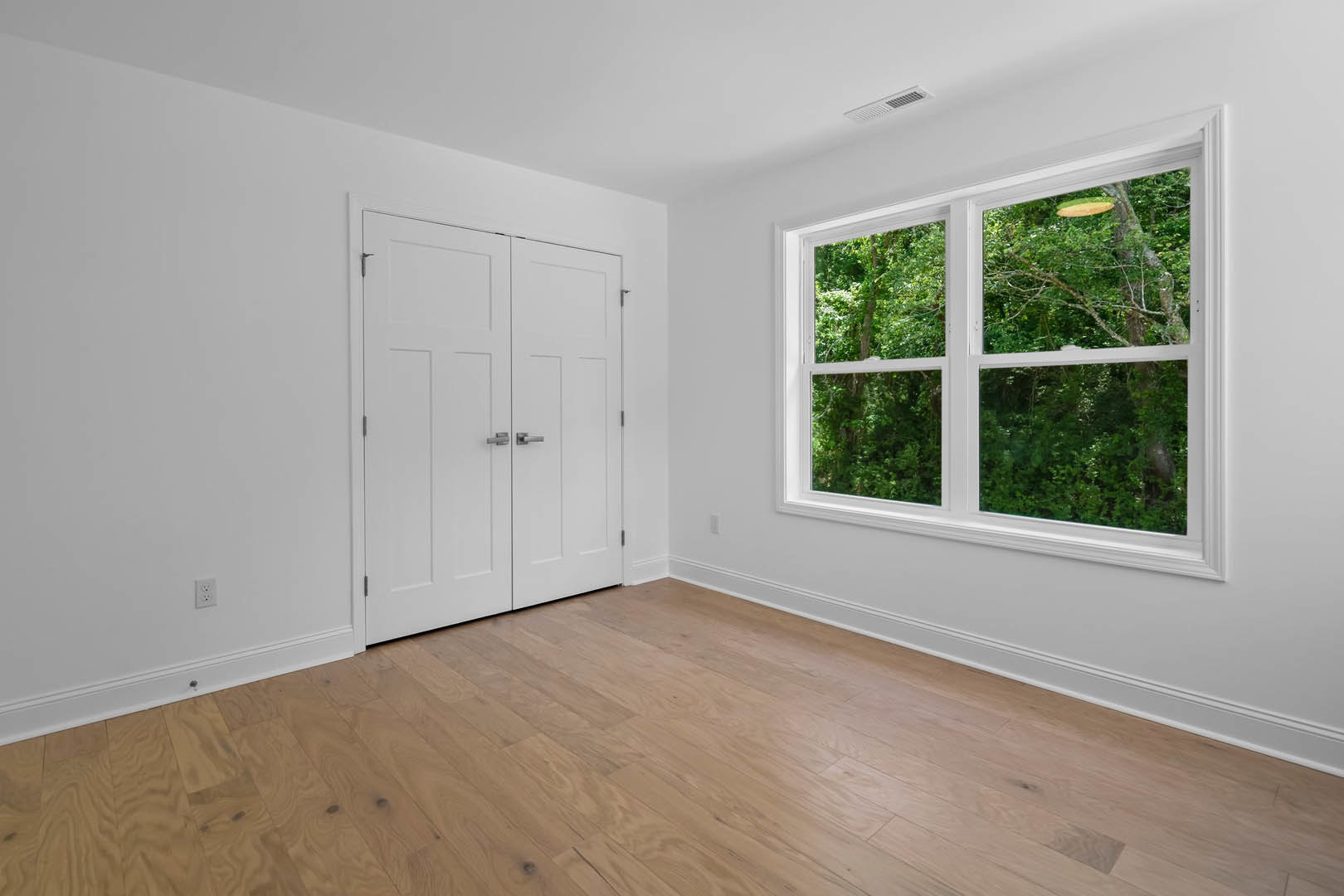 Wood floor and white walls in a room with a white door featuring silver handles, large window overlooking trees and forest.