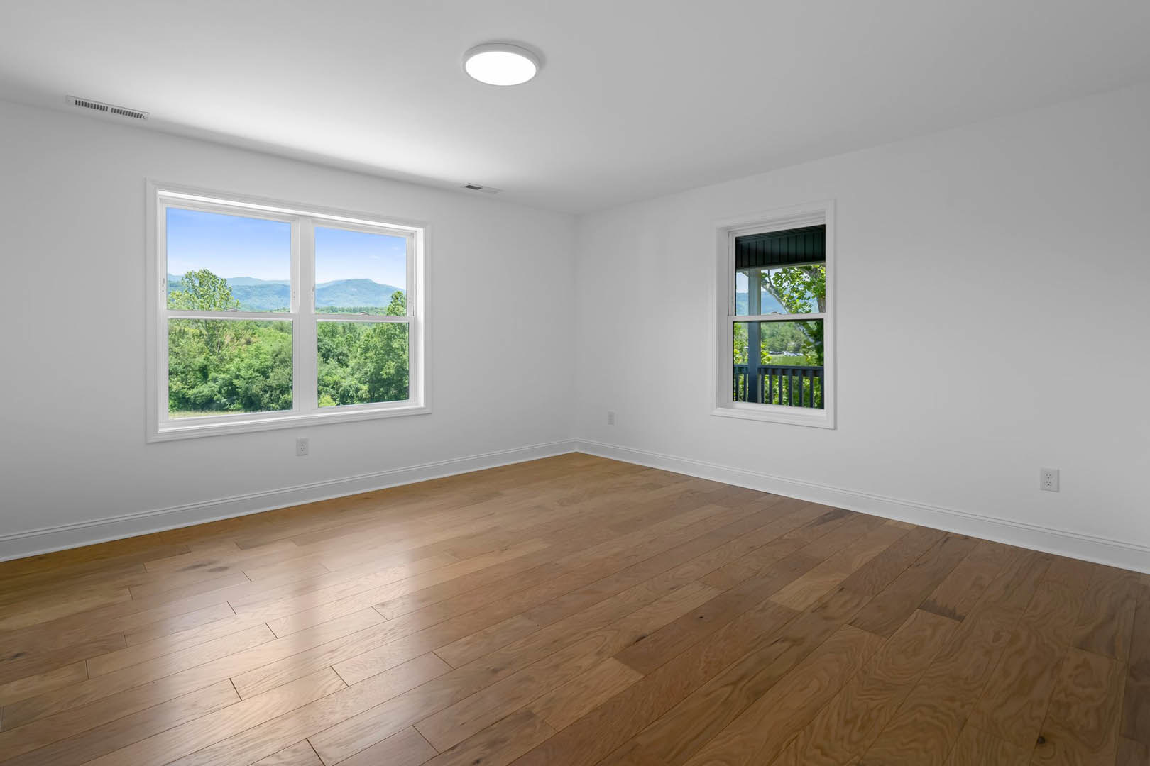 Spacious room featuring hardwood flooring, large windows with views of trees and distant mountains, white plaster walls, ceiling light fixture, and a close-up of a wood railing