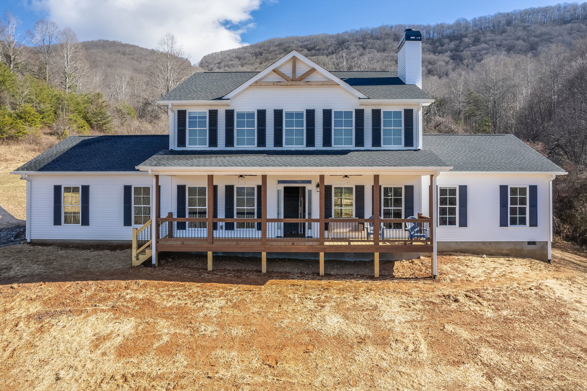 Two-story house with white-framed windows, wooden deck with railing, covered porch, grassy yard, and tree on a gentle hill under a cloudy sky