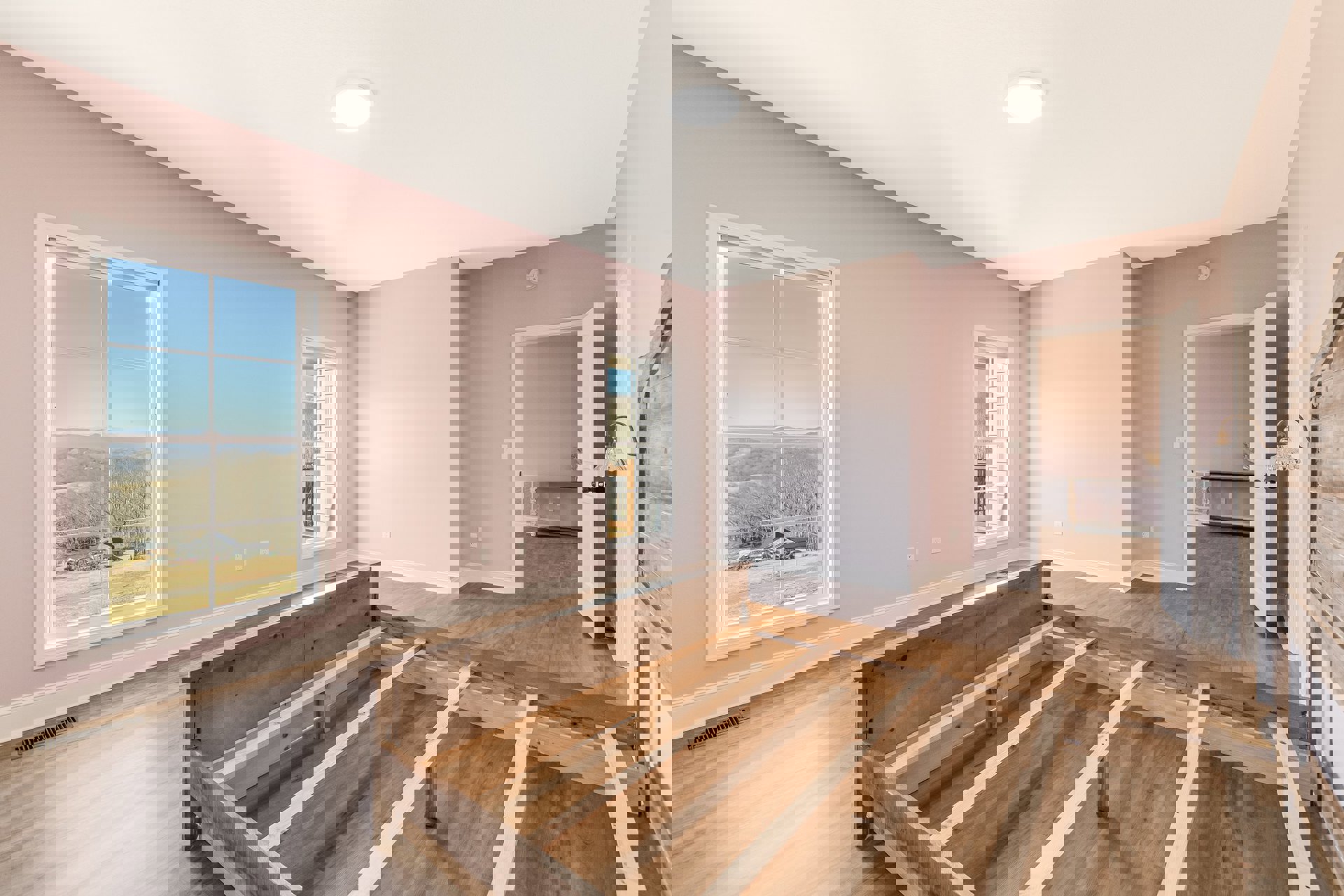 Bedroom with light wood flooring, white walls, bed frame, large window with white trim overlooking landscape, recessed ceiling light