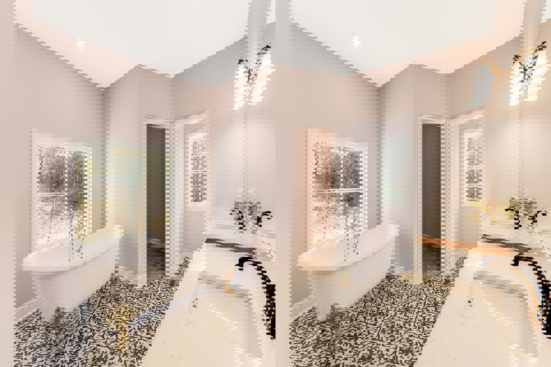 Bathroom with freestanding bathtub, white sink vanity, wall-mounted light fixture, large window overlooking trees and fence, gold decorative statue on patterned countertop, neutral