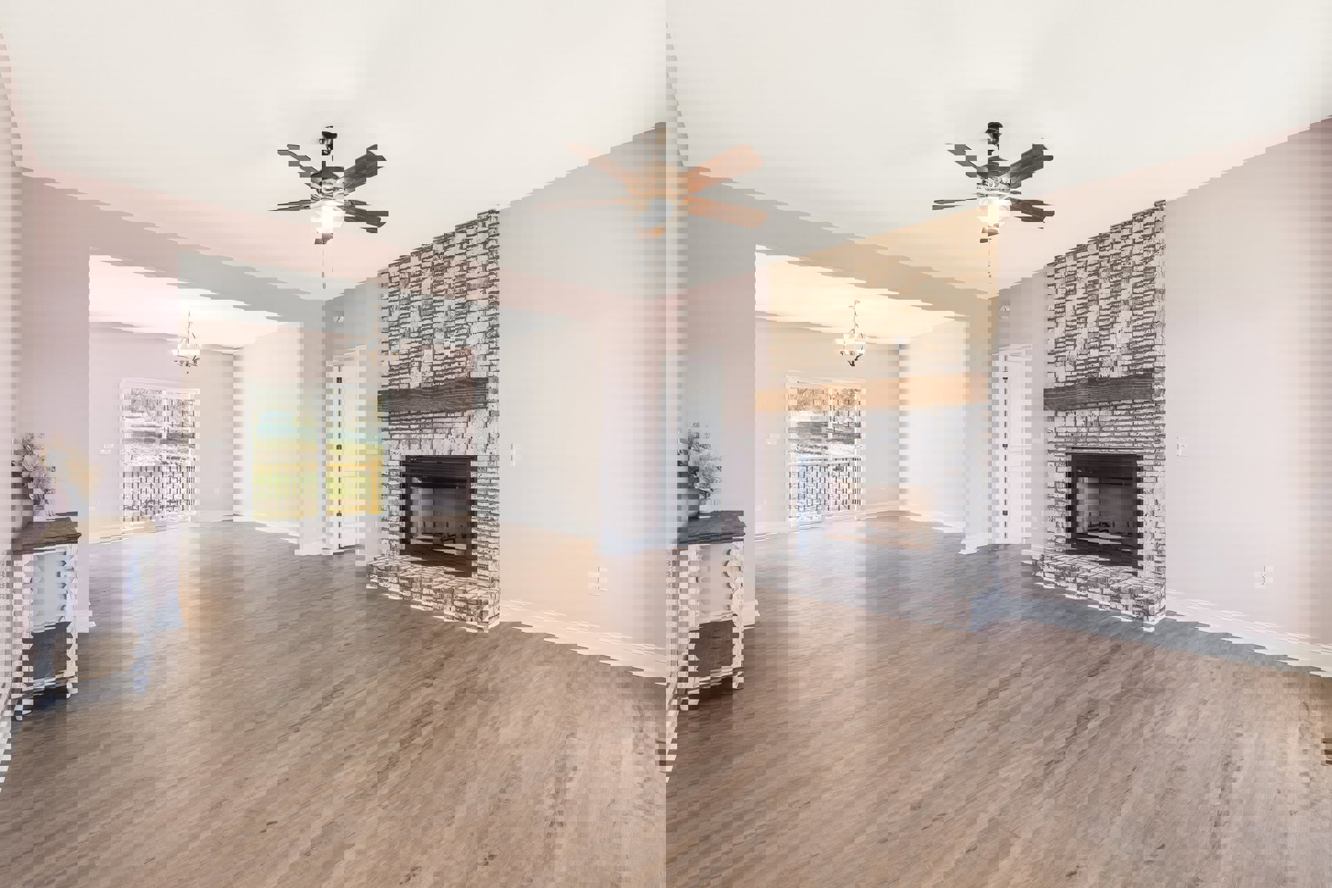 Living room with hardwood floors, central fireplace with glass door, ceiling fan with light, white table with wooden frame, sliding glass door leading to fenced outdoor area