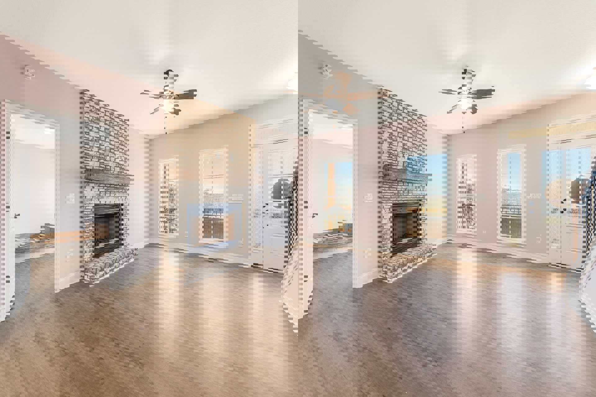 Living room with hardwood floors, ceiling fan with light, glass-door fireplace topped by a wood mantel shelf, large window offering outdoor view