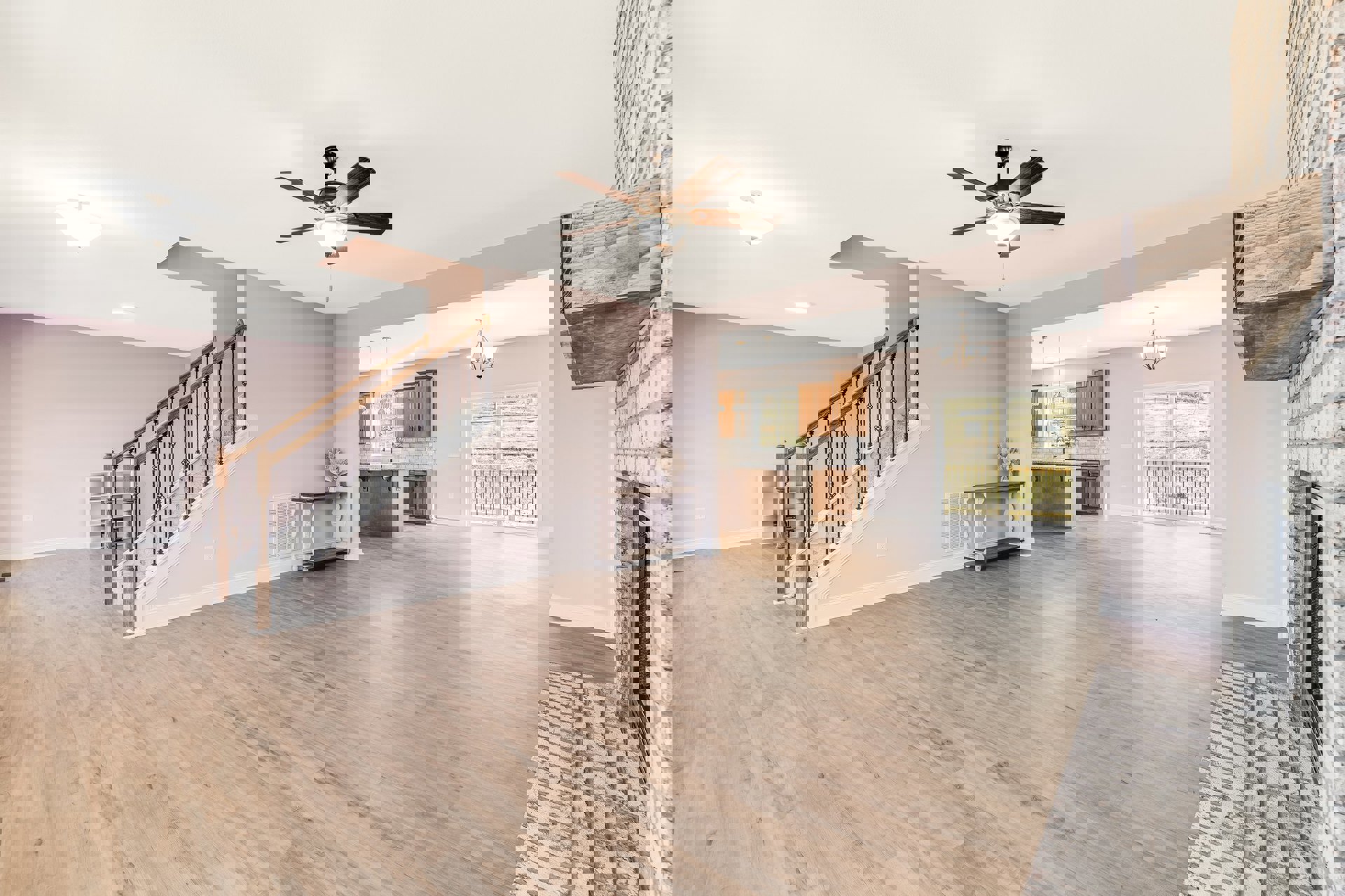 Spacious living area with wood flooring, central ceiling fan with light, staircase featuring wooden railing, large window overlooking fenced yard with grass, and a table beside a