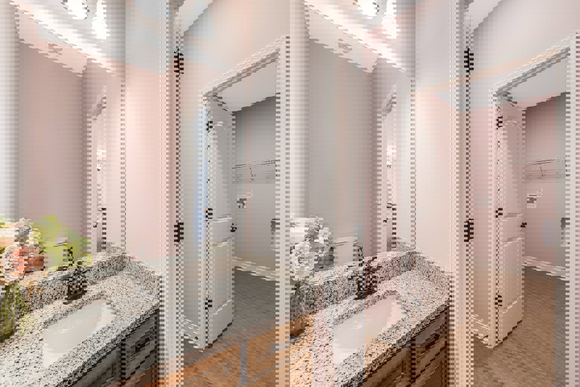 Marble countertop sink with chrome faucet, wood flooring, white wall-mounted light fixture, rectangular mirror, and vase of fresh flowers on vanity