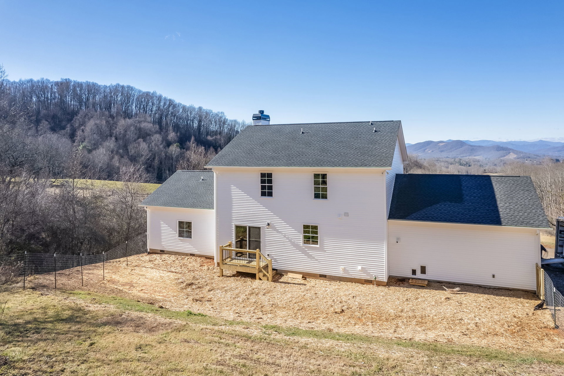 White house with black roof, wooden deck with railing, fenced yard, white-framed windows, grassy lawn, trees and mountains in background