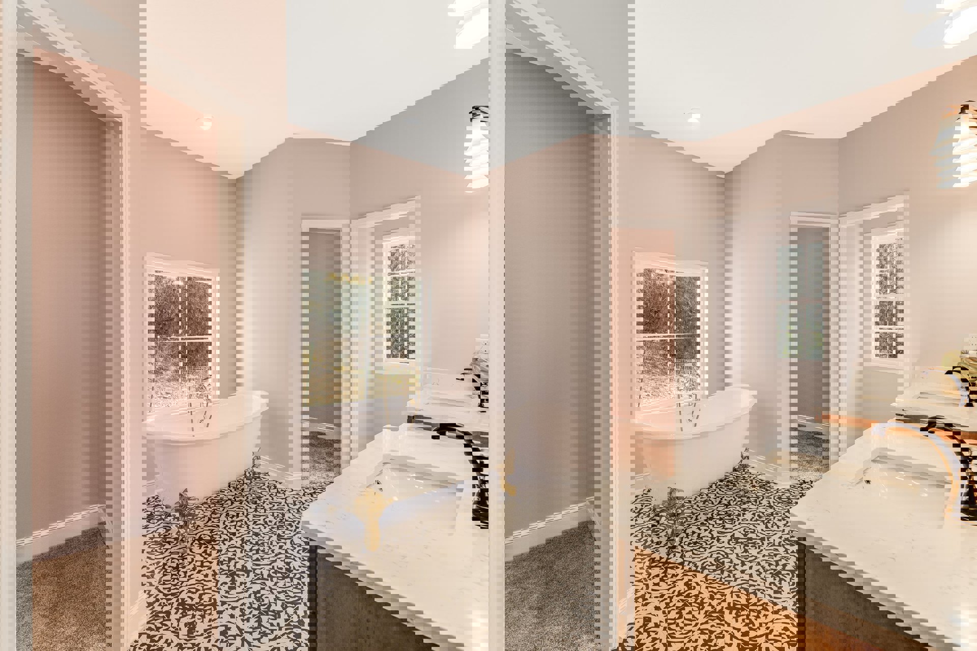 Freestanding white bathtub with gold legs beside a large window overlooking trees, marble tile floor, modern sink with gold faucet, and pendant light fixture