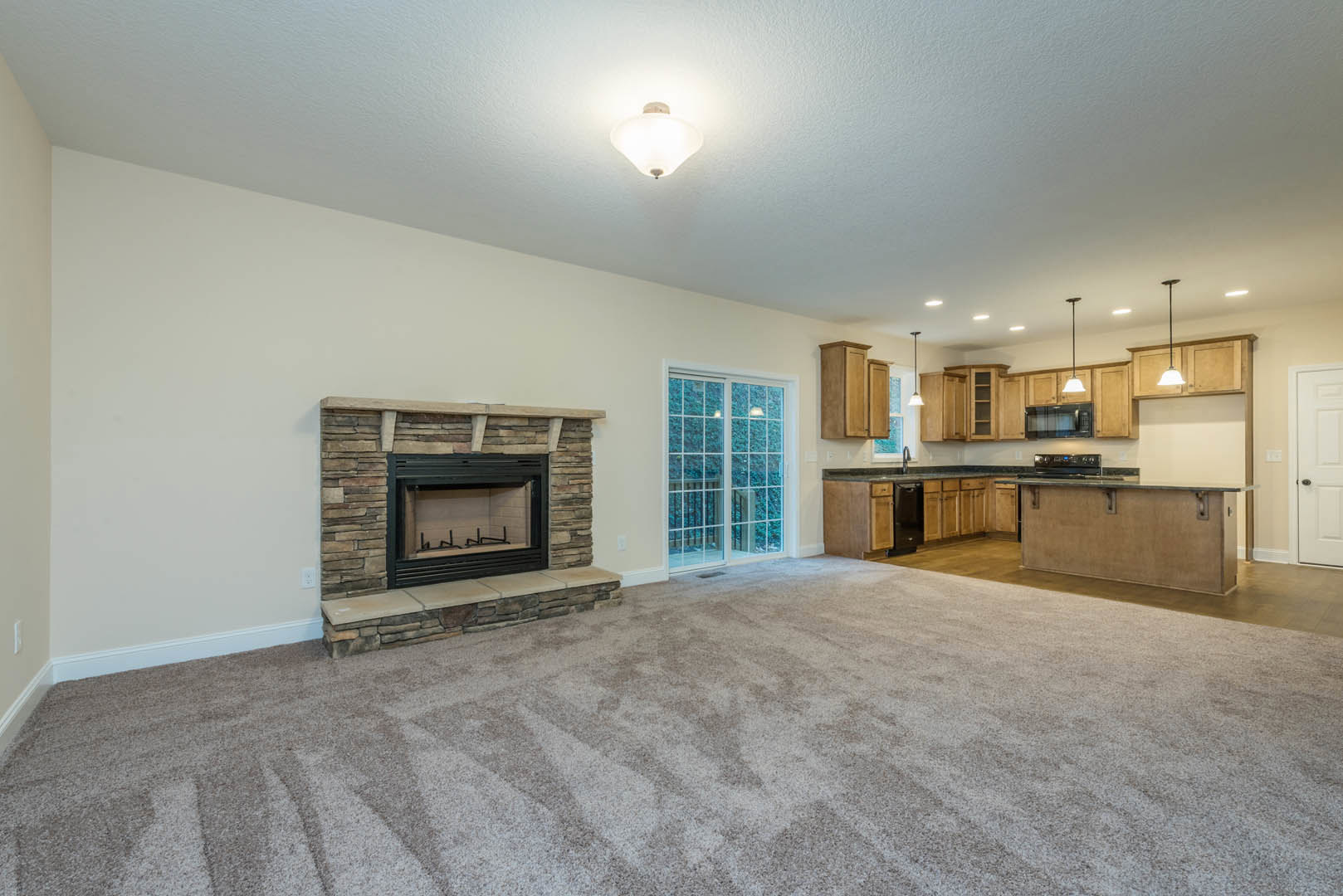 Open-concept living room with carpet flooring, stone fireplace featuring glass doors, view into kitchen with white cabinetry and pendant light fixtures, sliding glass door, and