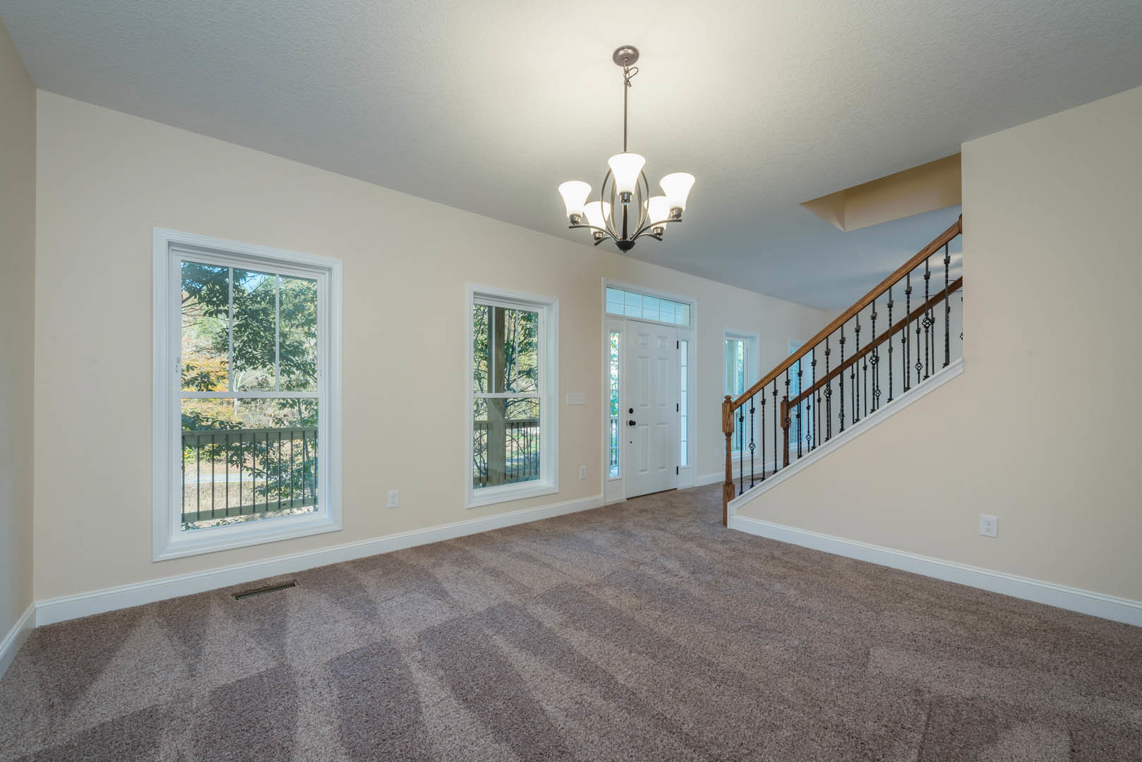 Carpeted living area featuring a staircase with metal railings, large window overlooking trees, and a modern chandelier hanging from a plaster ceiling with decorative molding.