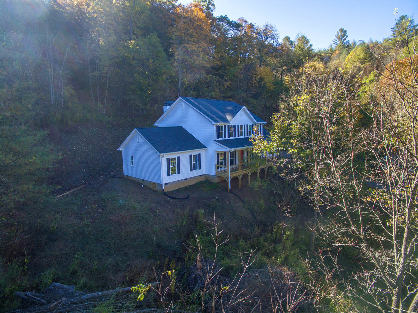 Modern farmhouse with black roof and white trim, elevated deck overlooking grassy hill, surrounded by dense woodland and tall trees under clear blue sky