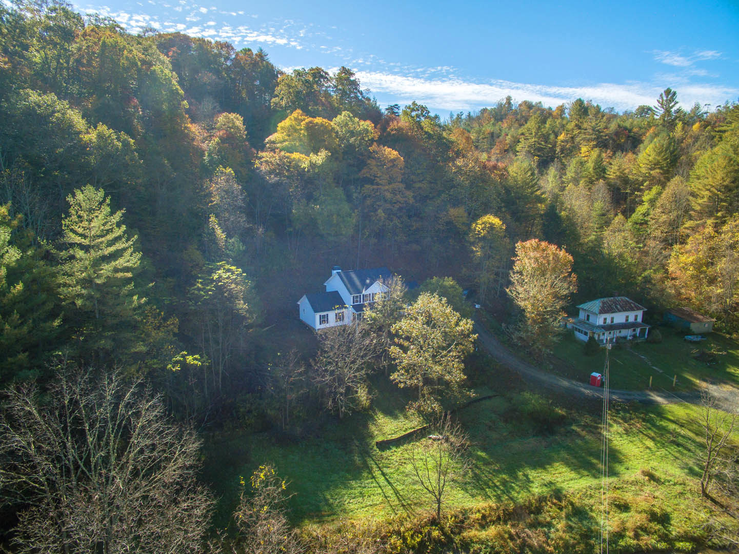 White house with green roof nestled among tall trees, autumn foliage, grassy lawn, blue sky with scattered clouds