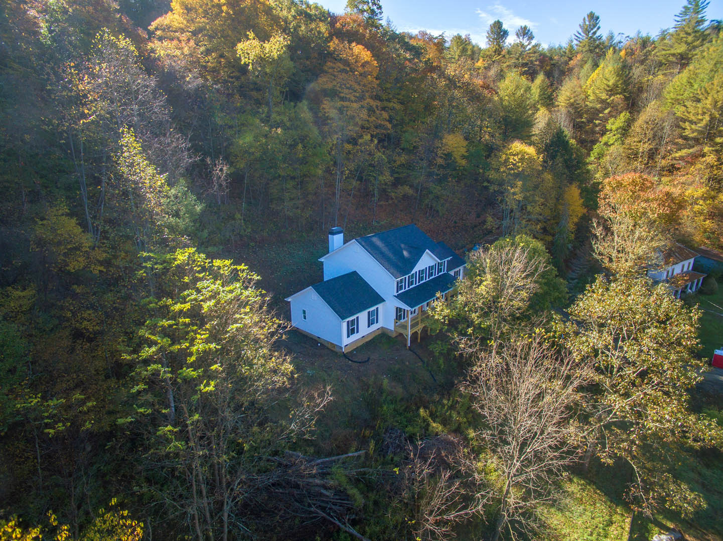 White house with black roof and covered front porch, nestled among tall green trees, surrounded by grass and natural landscaping under a blue sky.